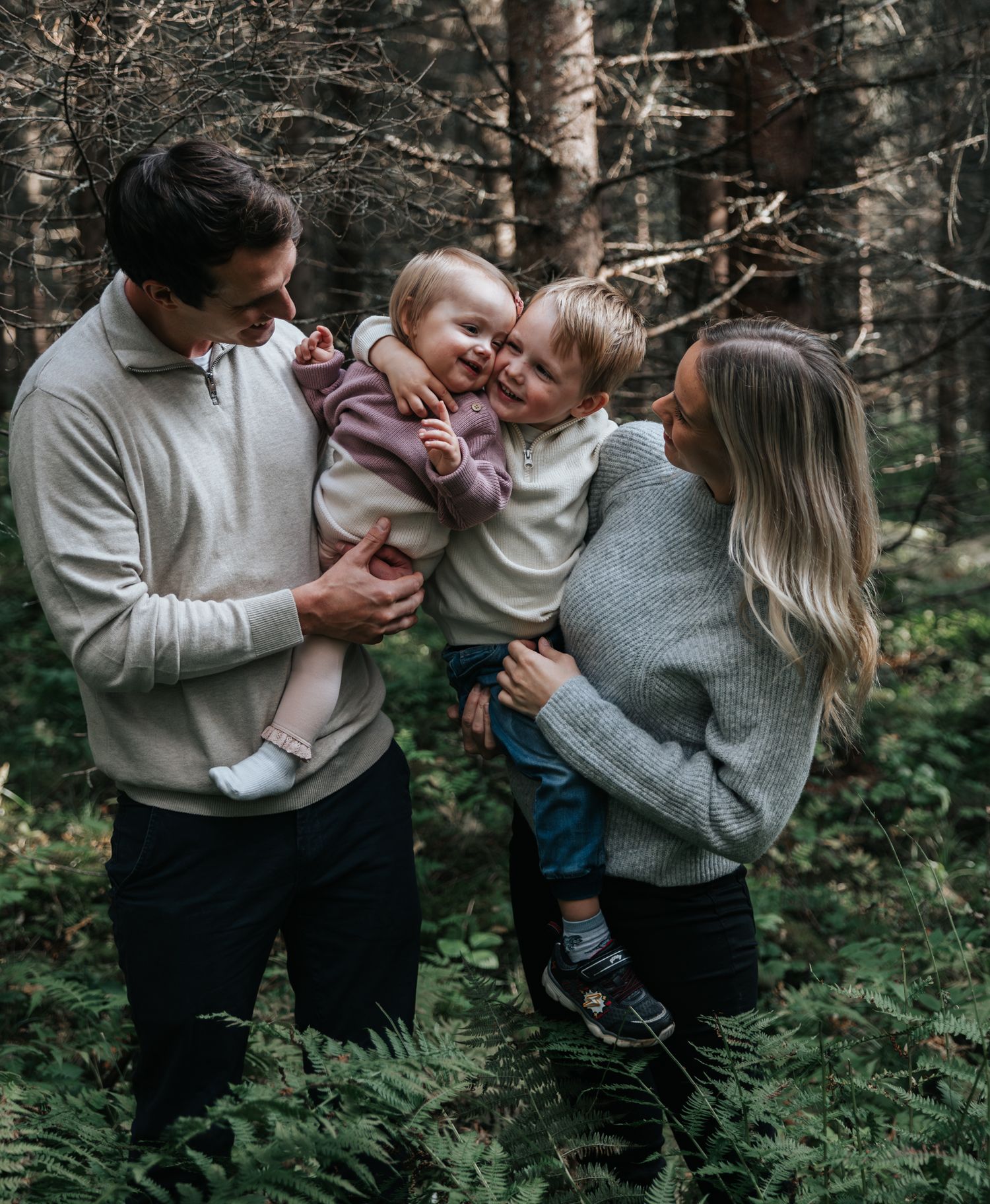 Parents holding two young children laughing together surrounded by ferns in a forest.
