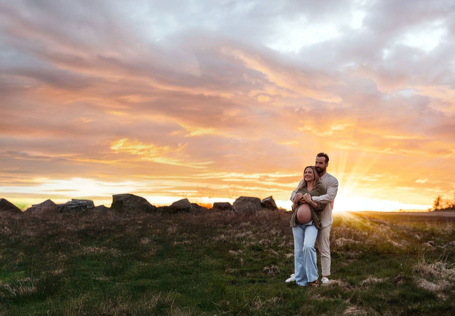 Couple embracing at golden sunset over rocky field, maternity photo shoot with vibrant orange and pink sky.