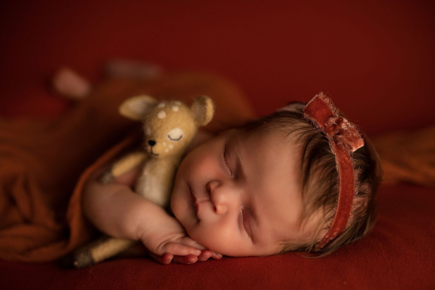 Newborn baby sleeping peacefully with stuffed toy on red backdrop wearing decorative headband.