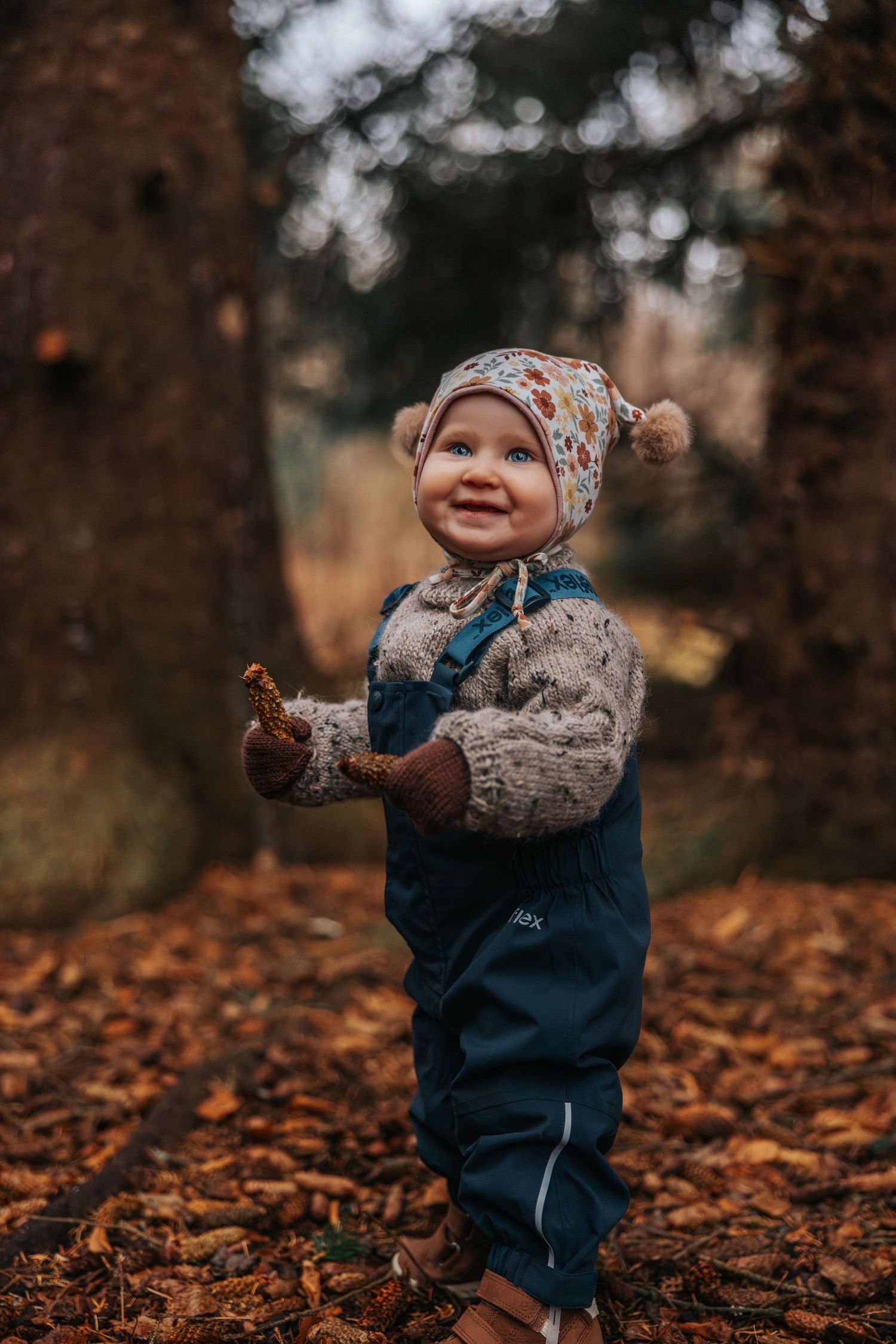 A smiling toddler in blue overalls, knit sweater, and floral bonnet plays outdoors among autumn leaves in a forest.