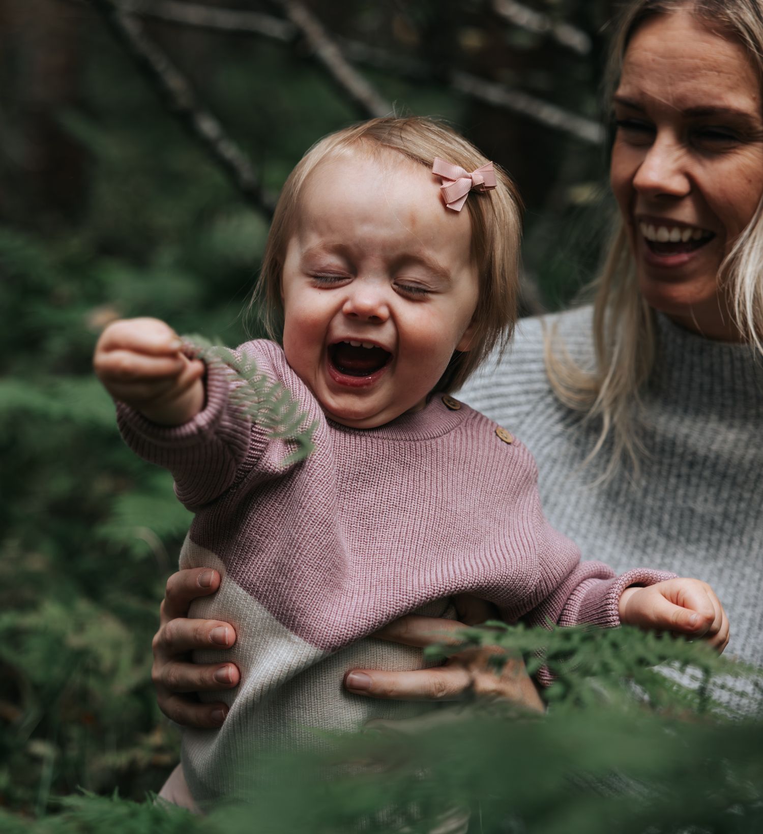 Laughing baby girl in pink knit sweater joyfully reaching out while mother smiles nearby.