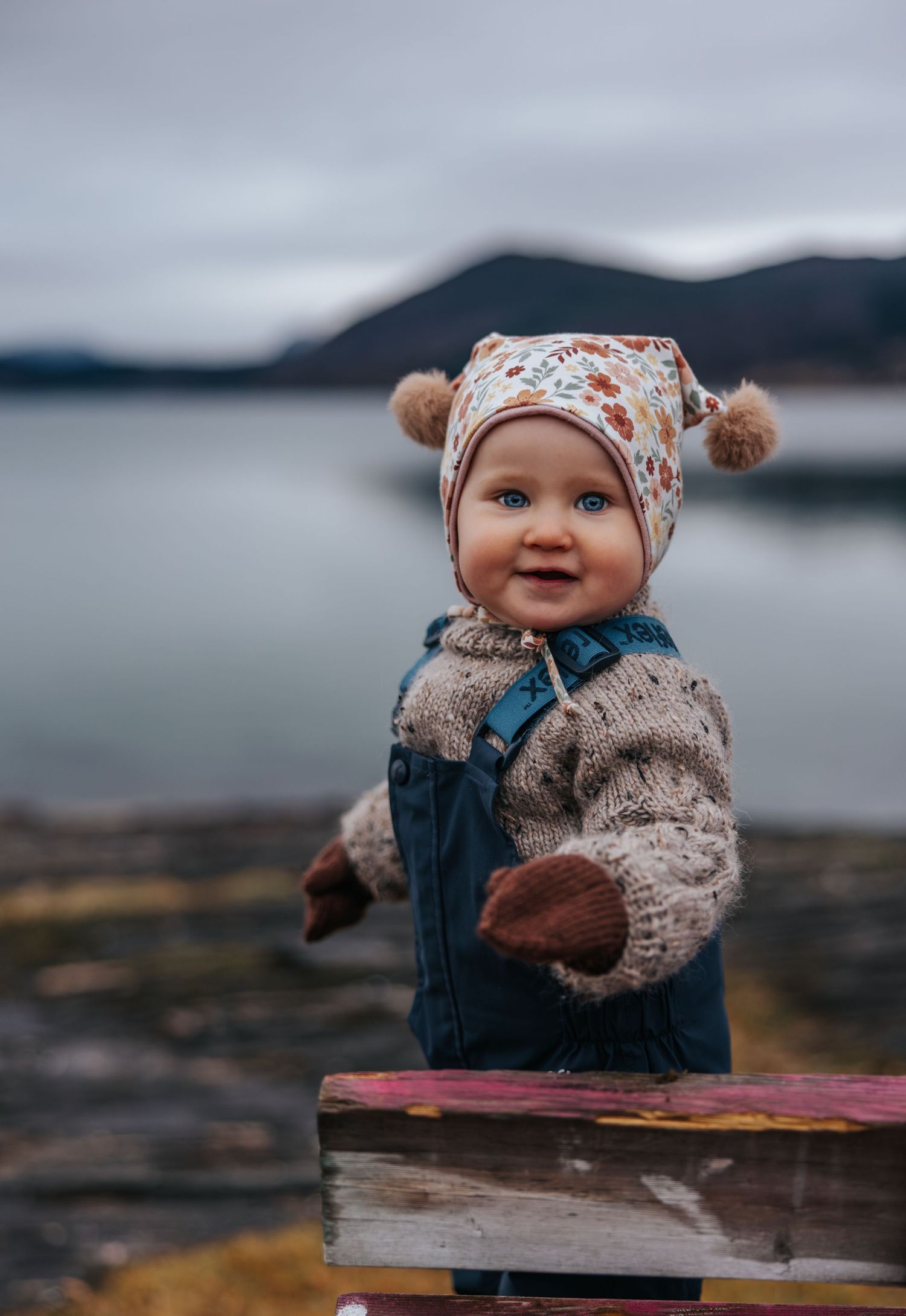 Smiling baby in floral hat and knit sweater outdoors near water.