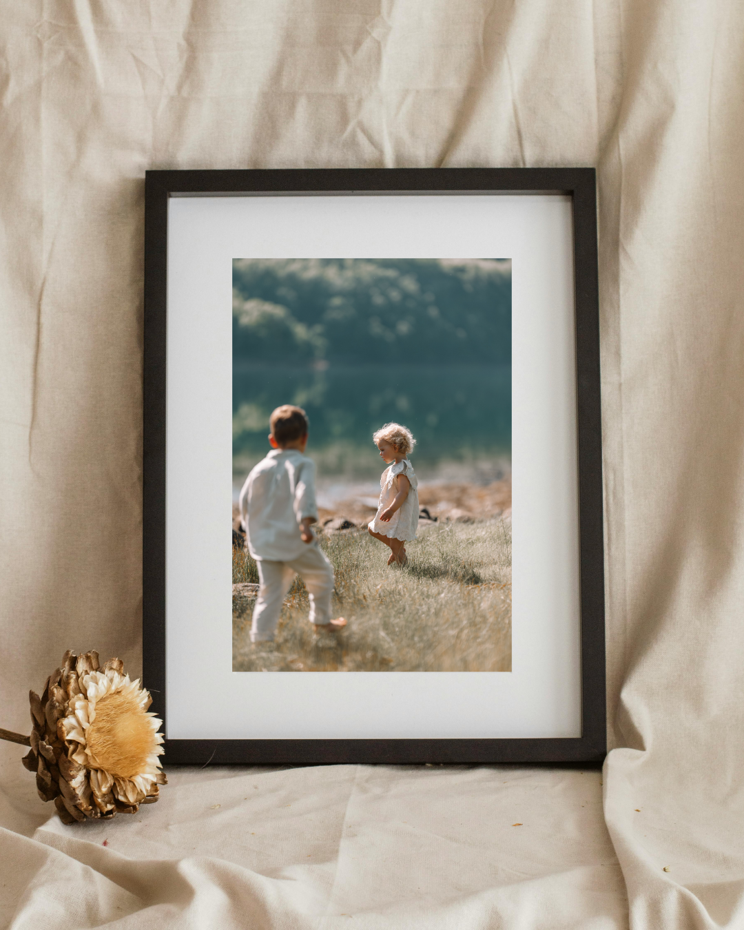 Framed photo of two young children playing in a meadow near a lake, displayed on linen fabric.