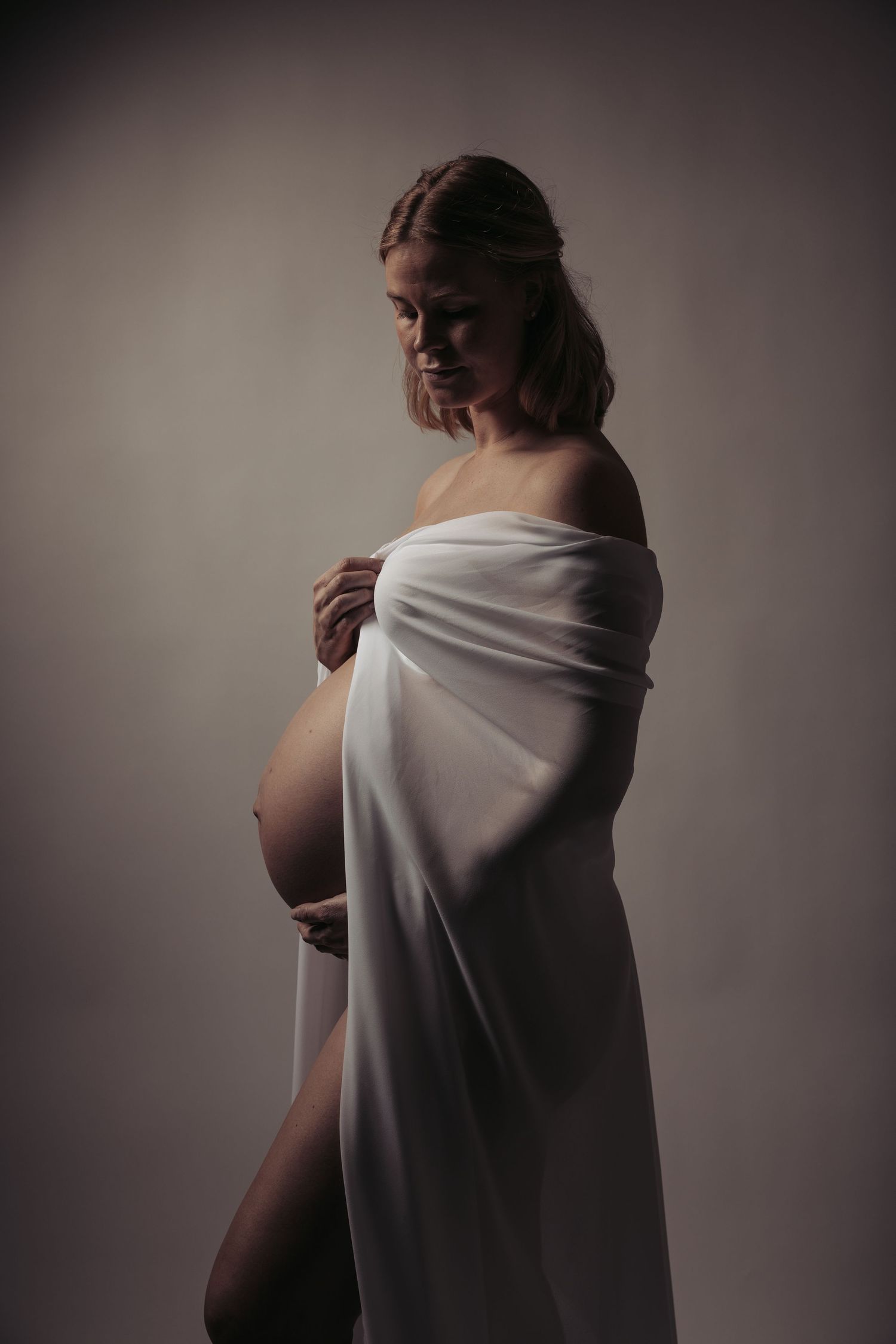 Pregnant woman draped in white fabric, cradling bump in moody studio portrait.