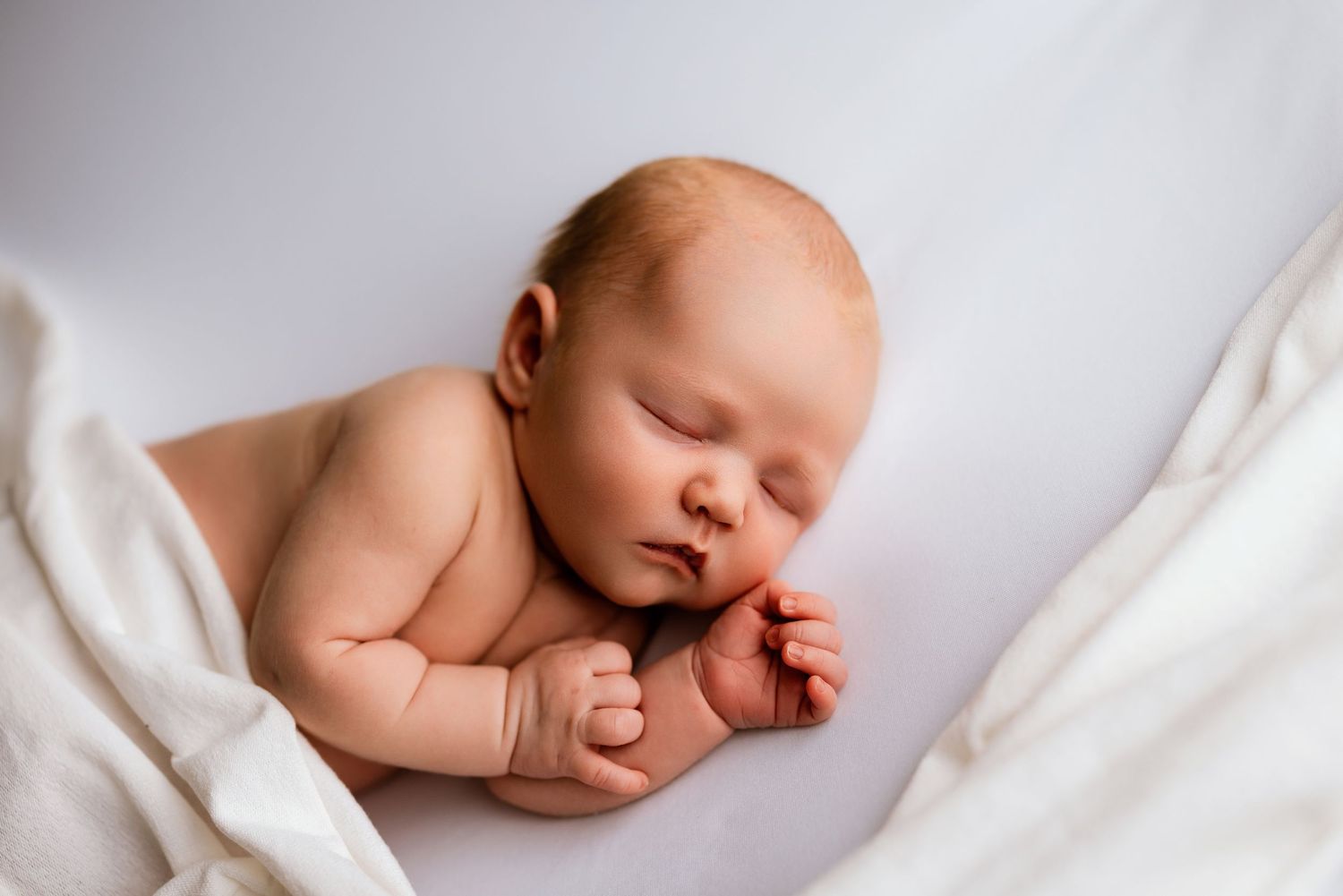 Peaceful newborn baby sleeping on white blanket, curled up with tiny fists, soft natural light photography.