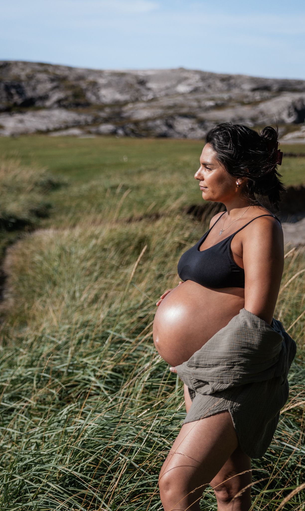 Pregnant woman in black sports bra holding baby bump in lush green coastal meadow.