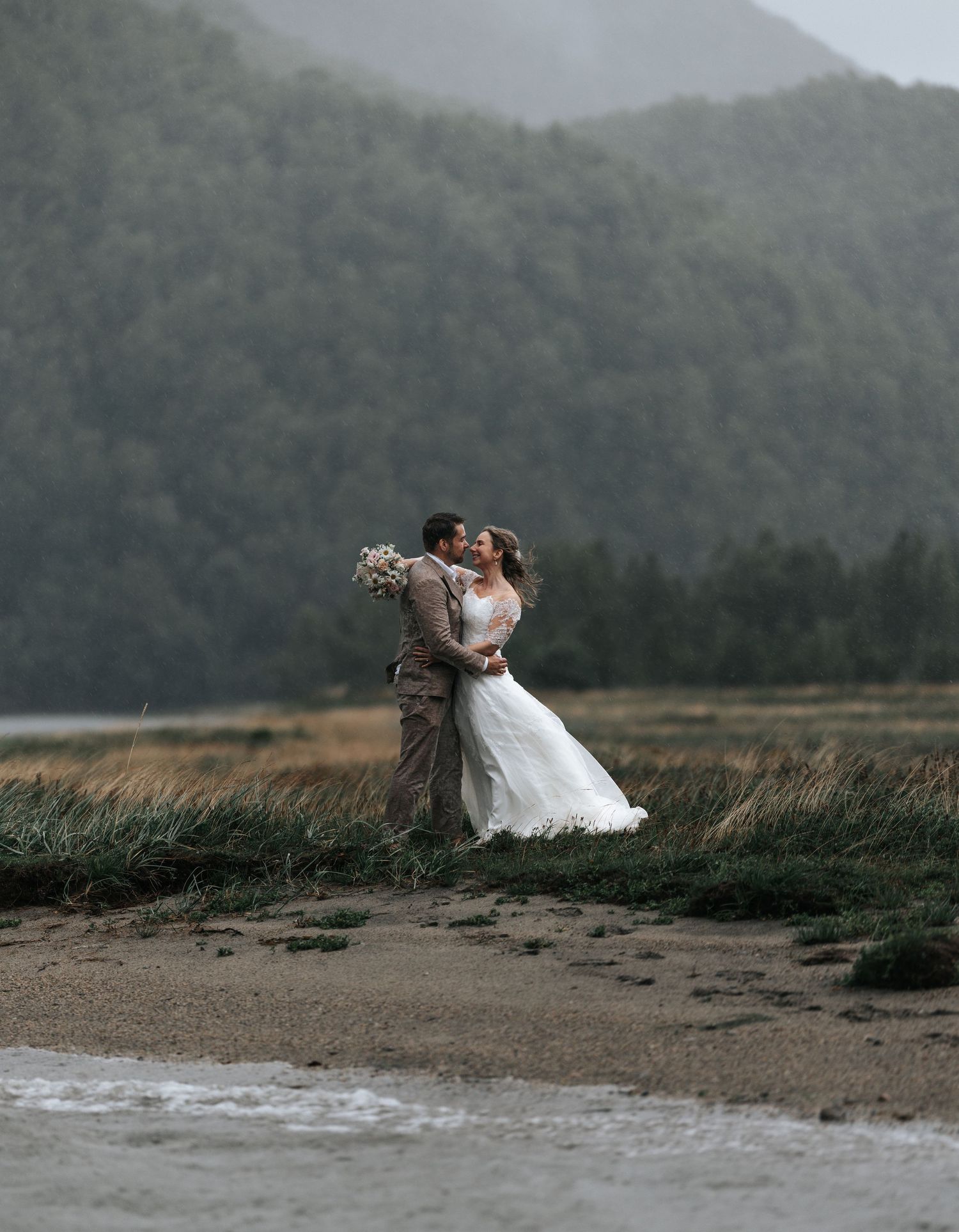 Bride and groom sharing a kiss on a sandy beach with misty mountains in the background.