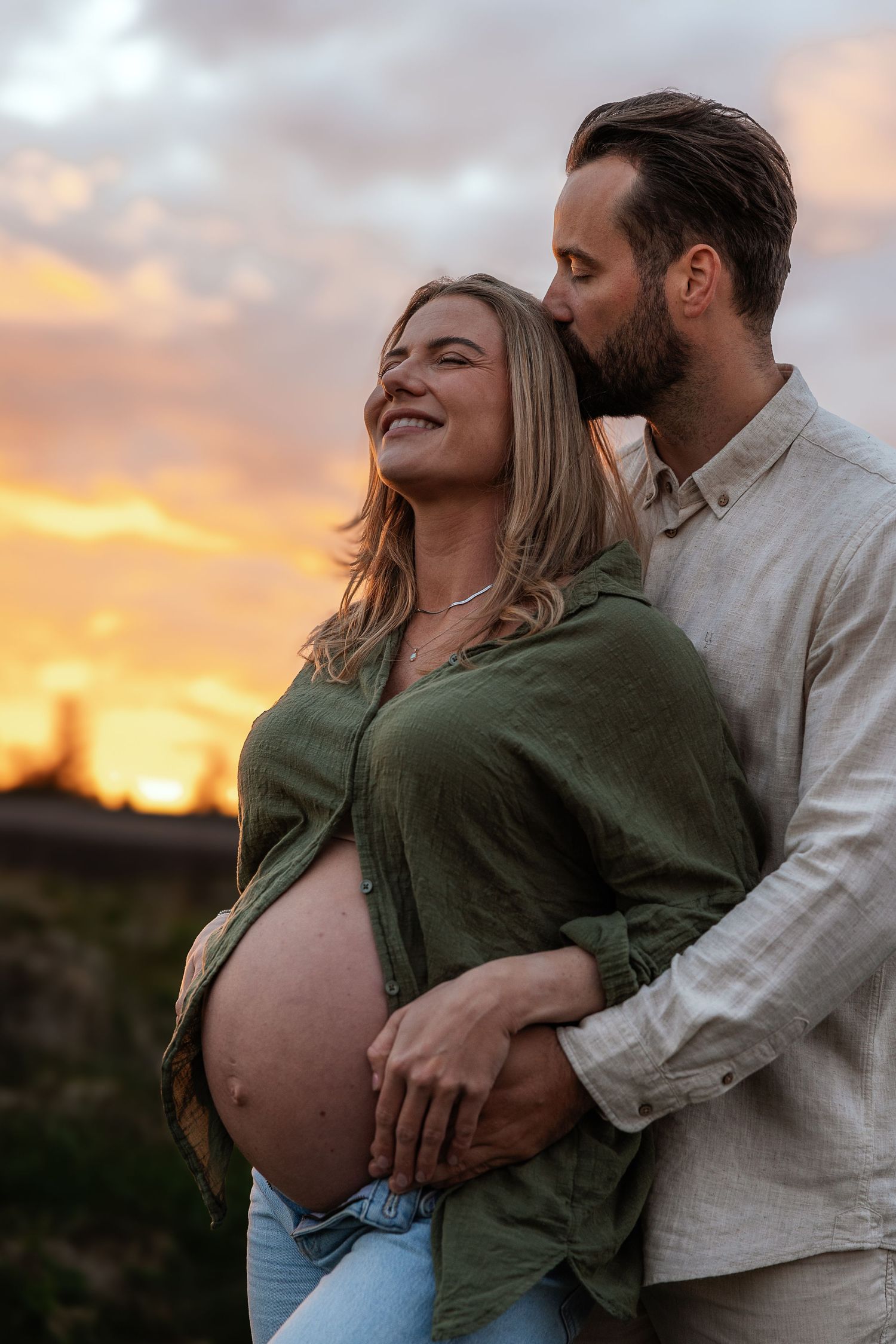 A couple shares a tender sunset moment, man kissing pregnant woman's head while cradling her baby bump outdoors.