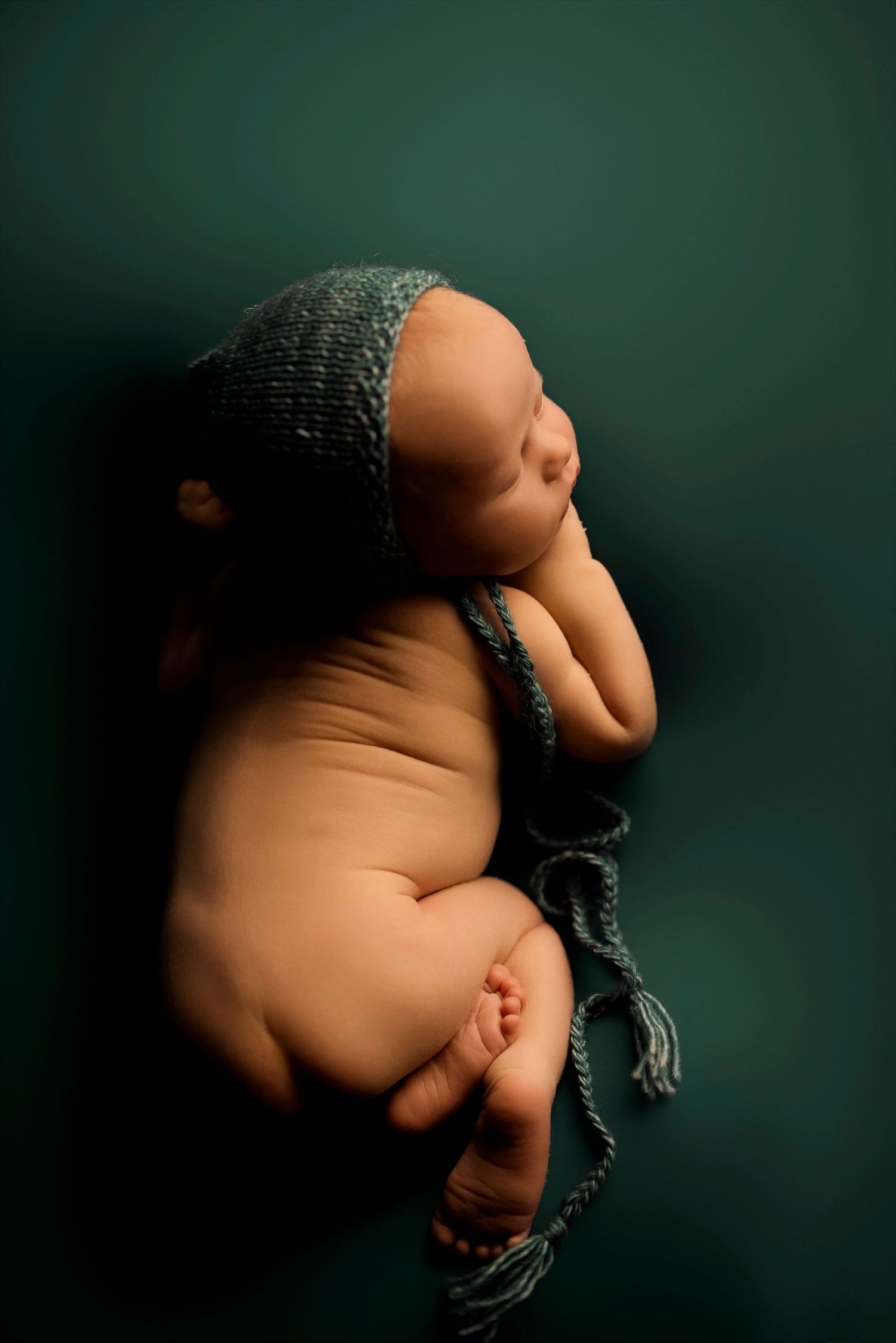 Newborn baby wearing dark knit bonnet sleeping peacefully on green backdrop in classic portrait pose.