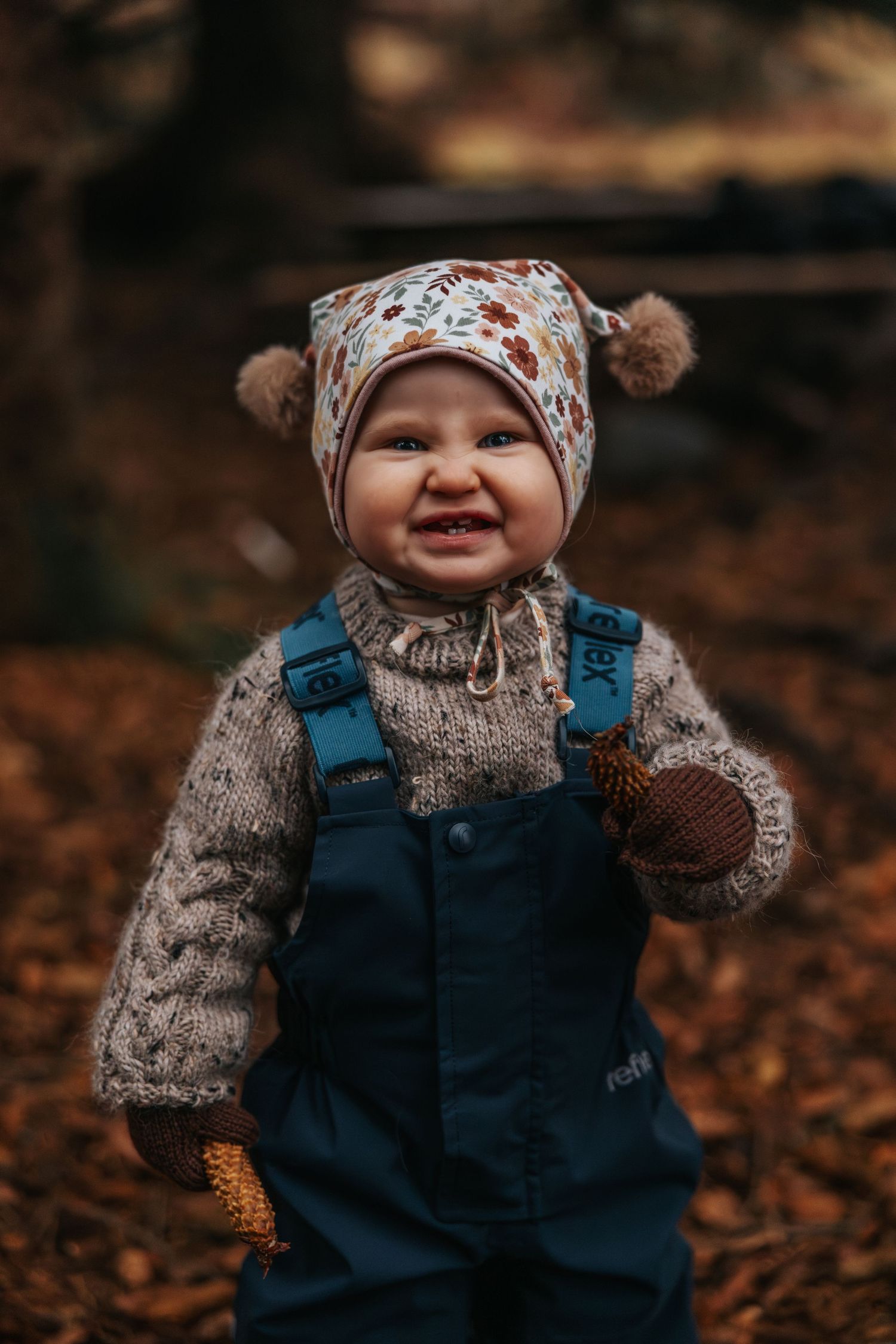 Happy baby in blue overalls and floral hat standing in autumn leaves.