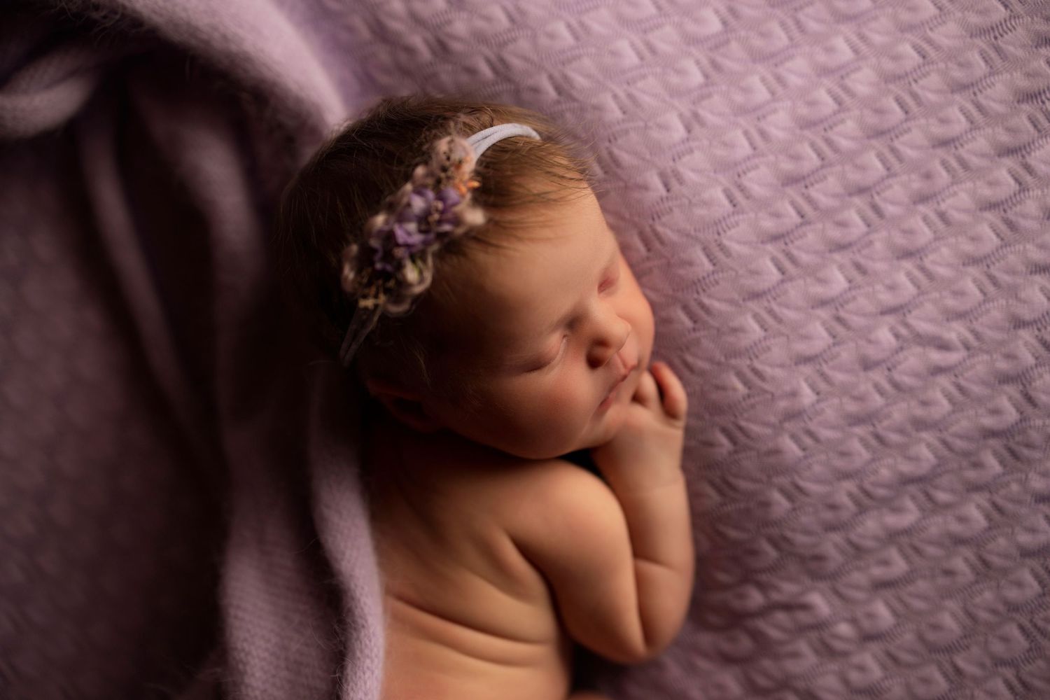 Newborn baby sleeping peacefully on purple textured blanket wearing floral headband in professional portrait.
