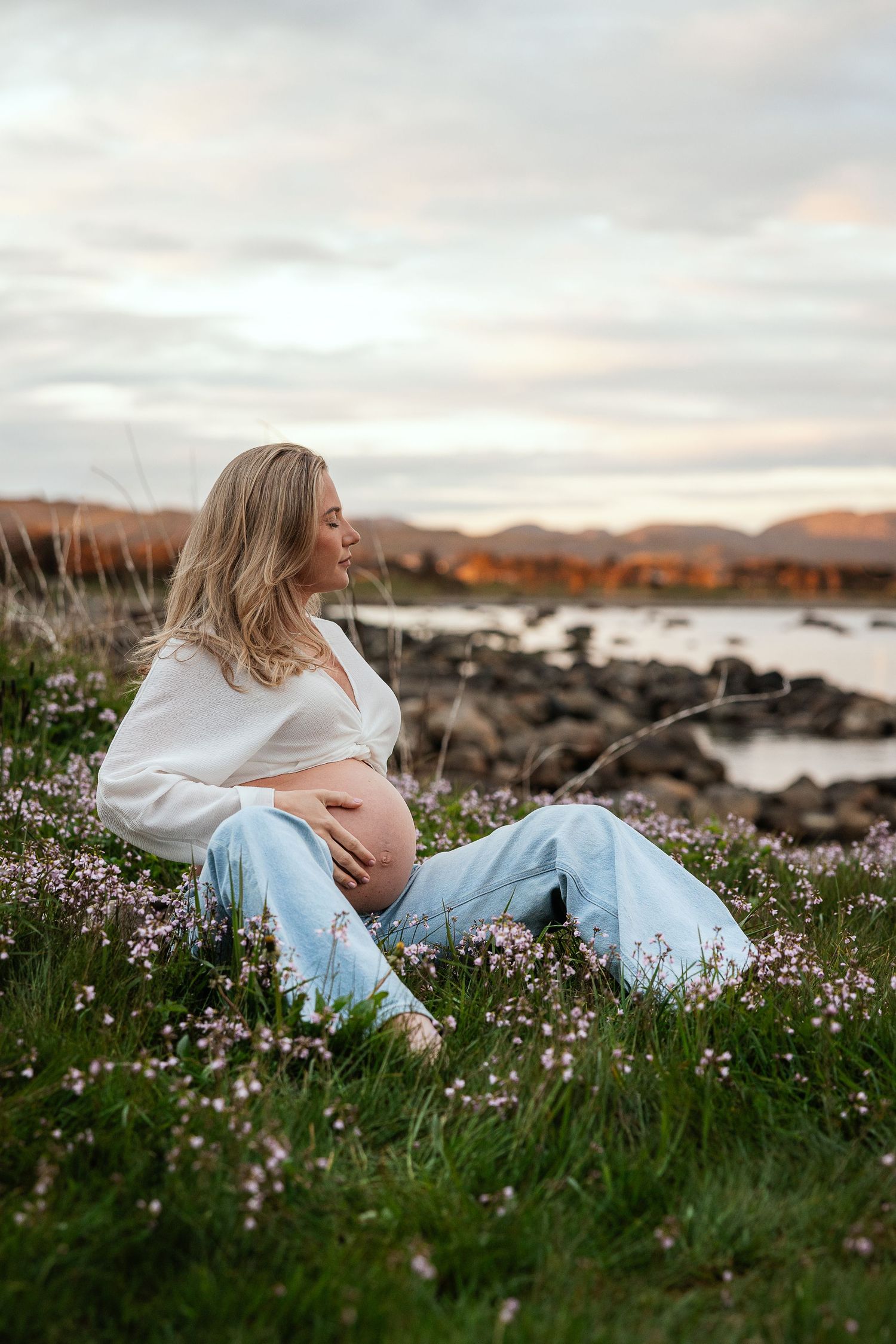 Pregnant woman sitting in wildflowers by rocky shore at sunset, cradling baby bump in white top and jeans.