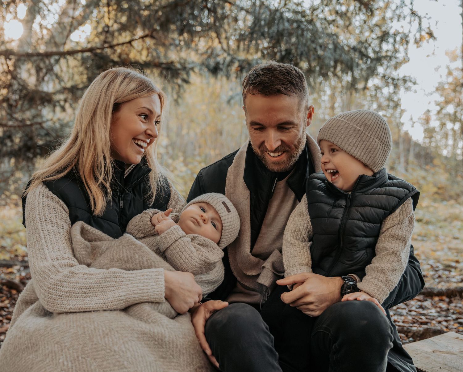 A happy family of four sitting outdoors in fall, parents smiling with newborn and young child.
