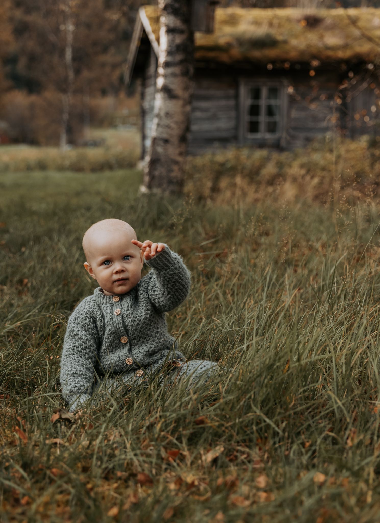 A smiling baby in a gray knitted sweater sits in tall grass near a rustic wooden cabin in an autumn field.