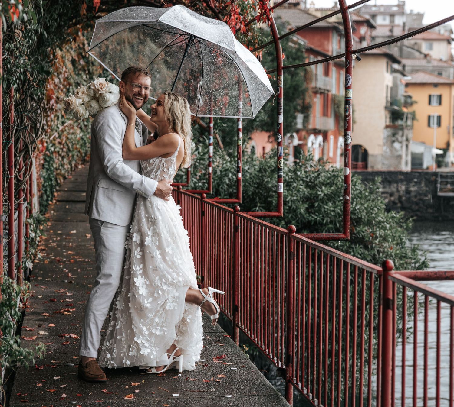 Couple embracing under clear umbrella on rainy bridge walkway, bride in floral lace gown, colorful backdrop.