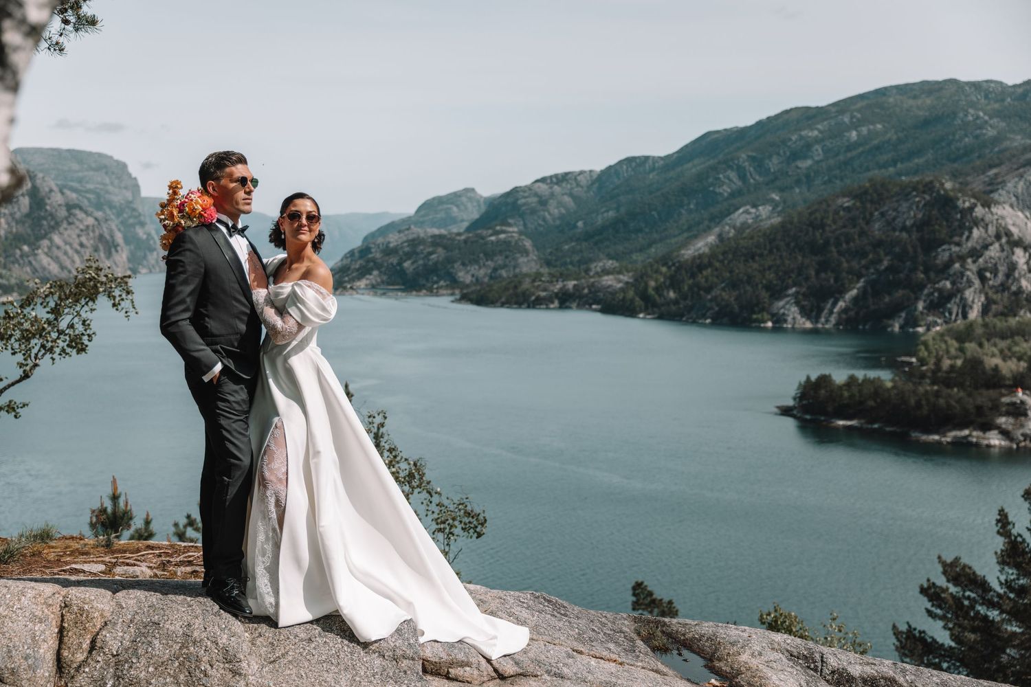 Bride and groom standing on rocky cliff overlooking scenic Norwegian fjord landscape on wedding day.