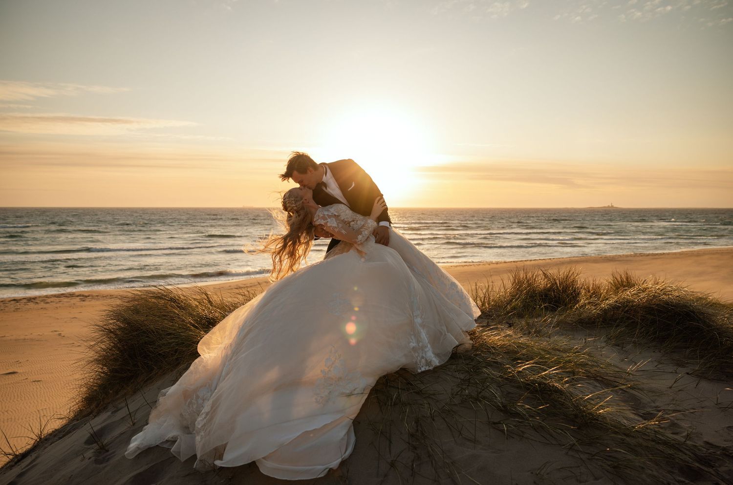 Groom dipping bride in voluminous white gown on sand dunes at breathtaking golden sunset beach wedding.