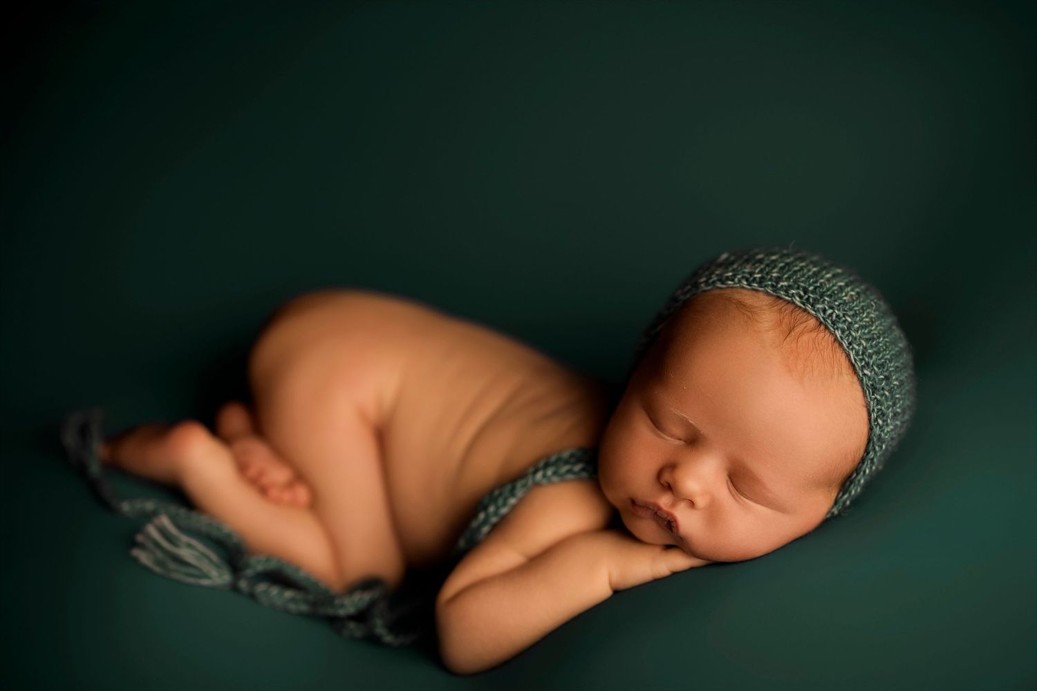 Peaceful newborn baby sleeping curled up wearing a teal knit hat on a dark green backdrop.