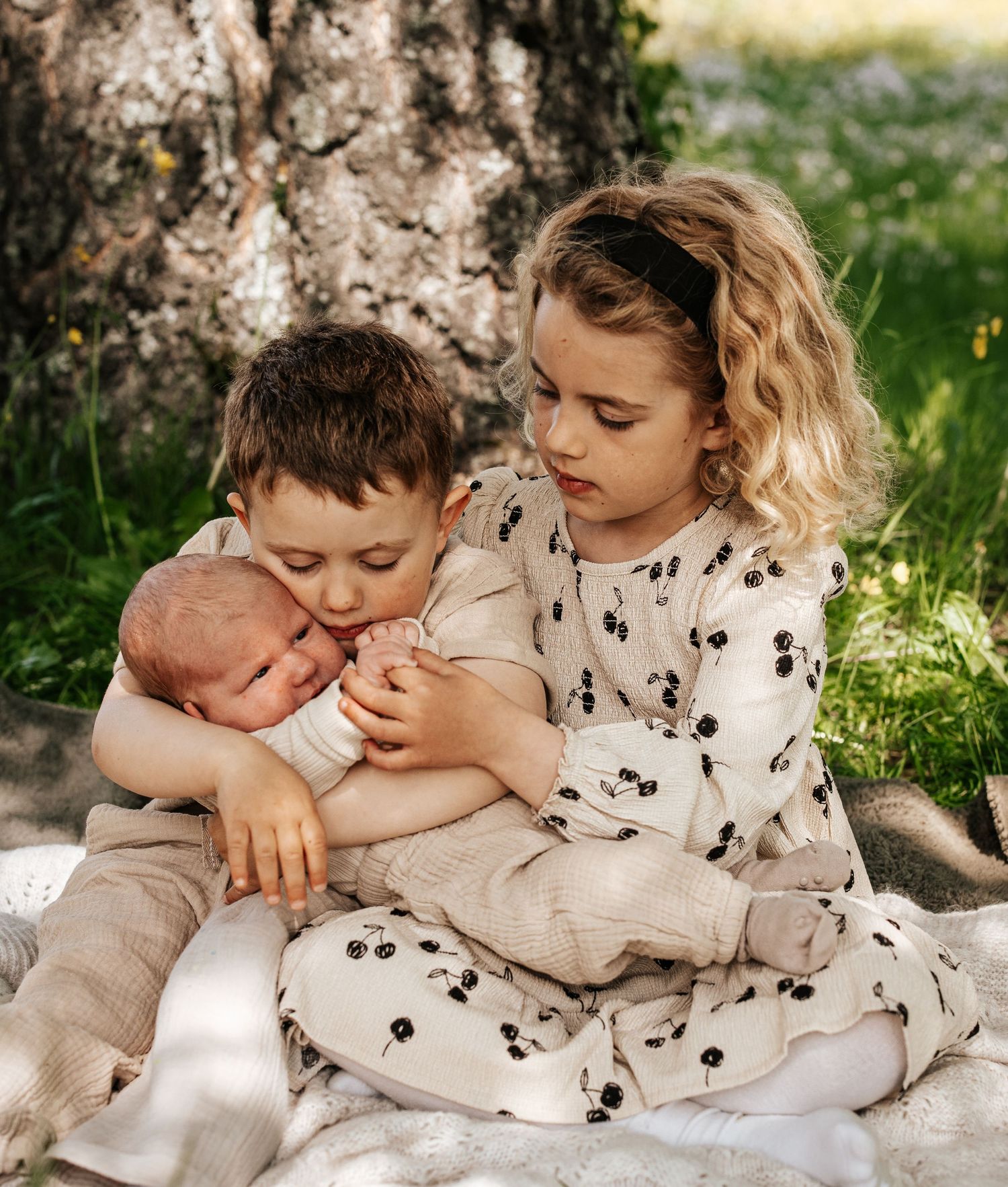 Three siblings gently holding newborn baby outdoors under a tree.