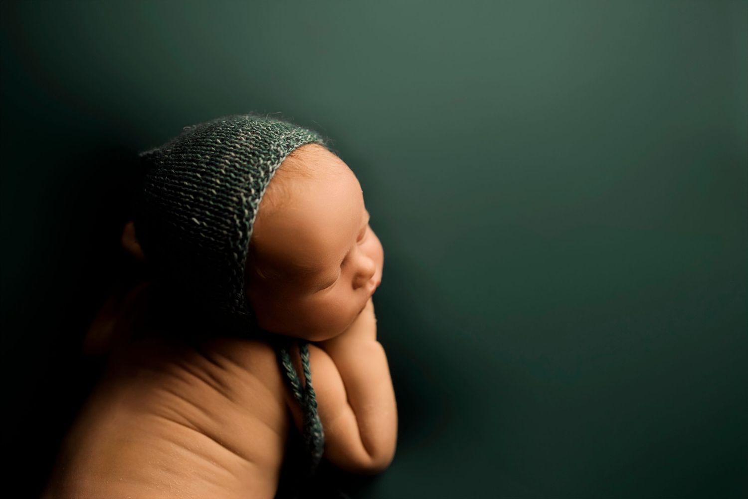 Newborn baby doll wearing a knitted dark green bonnet against a matching green background in a minimalist portrait.