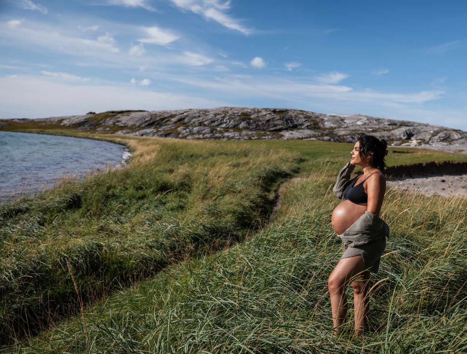 Pregnant woman standing in tall grass by a rocky coastline under a blue sky.