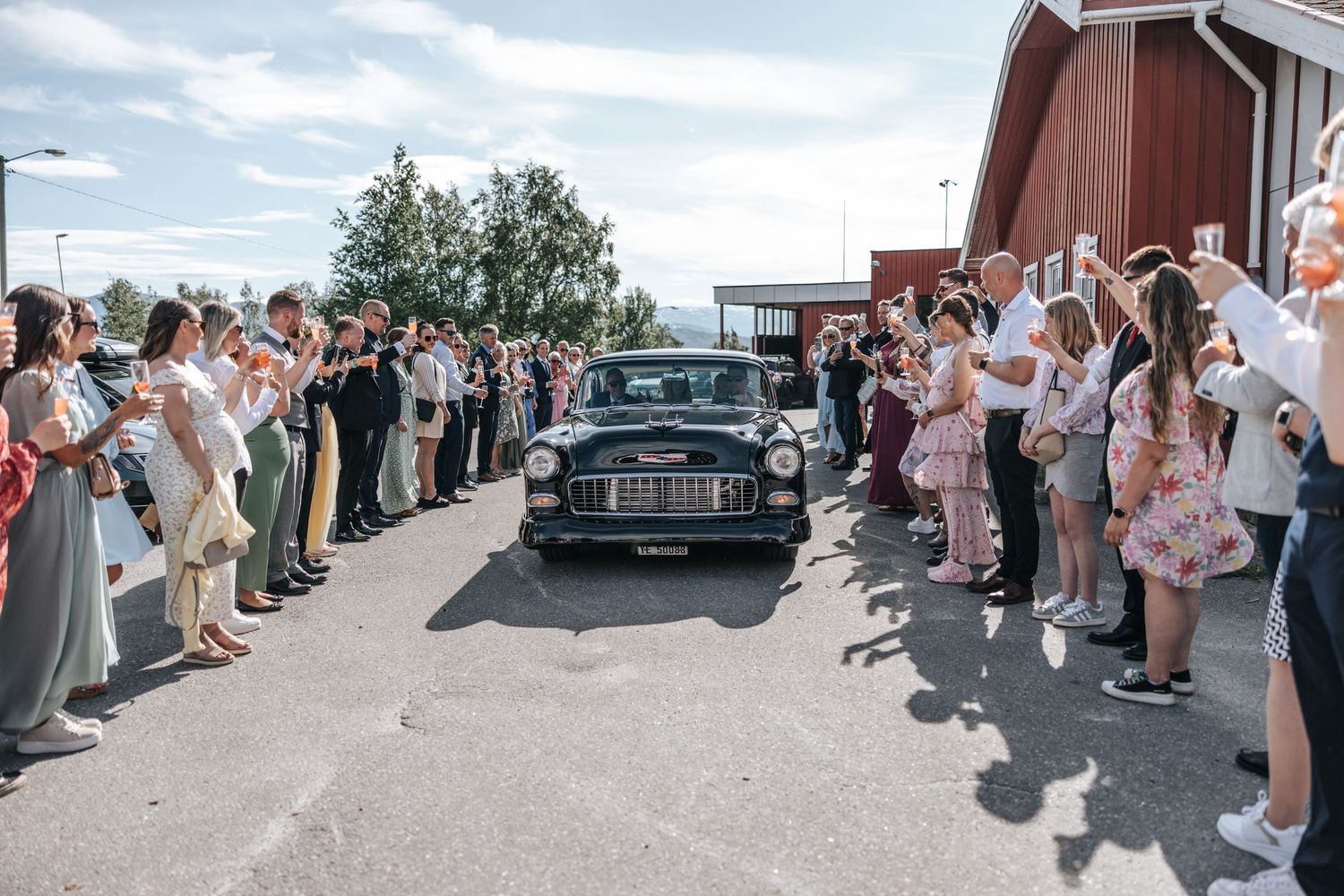 Classic black vintage car driving between rows of cheering wedding guests outside a red barn.