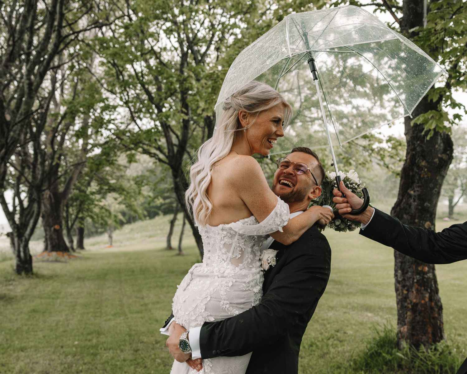 Groom lifting bride in white lace dress under clear umbrella in rainy outdoor wedding setting.