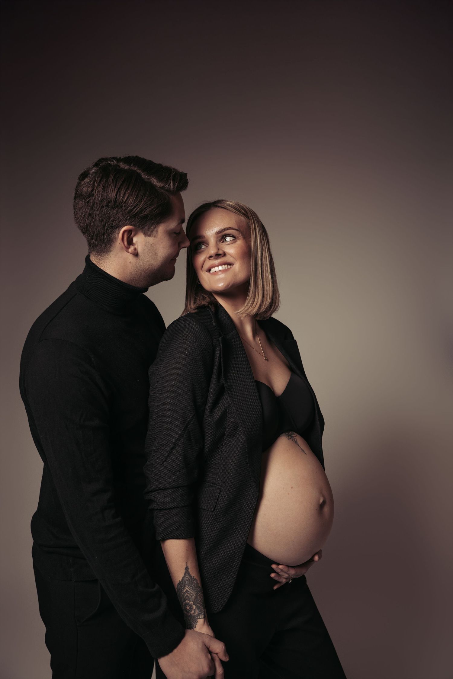 Pregnant woman in black blazer smiling at partner during maternity photo shoot against dark background.