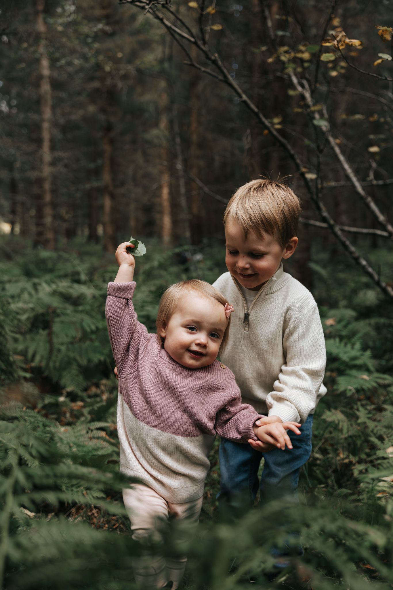 Toddler siblings in coordinating outfits smiling together among lush green forest ferns.