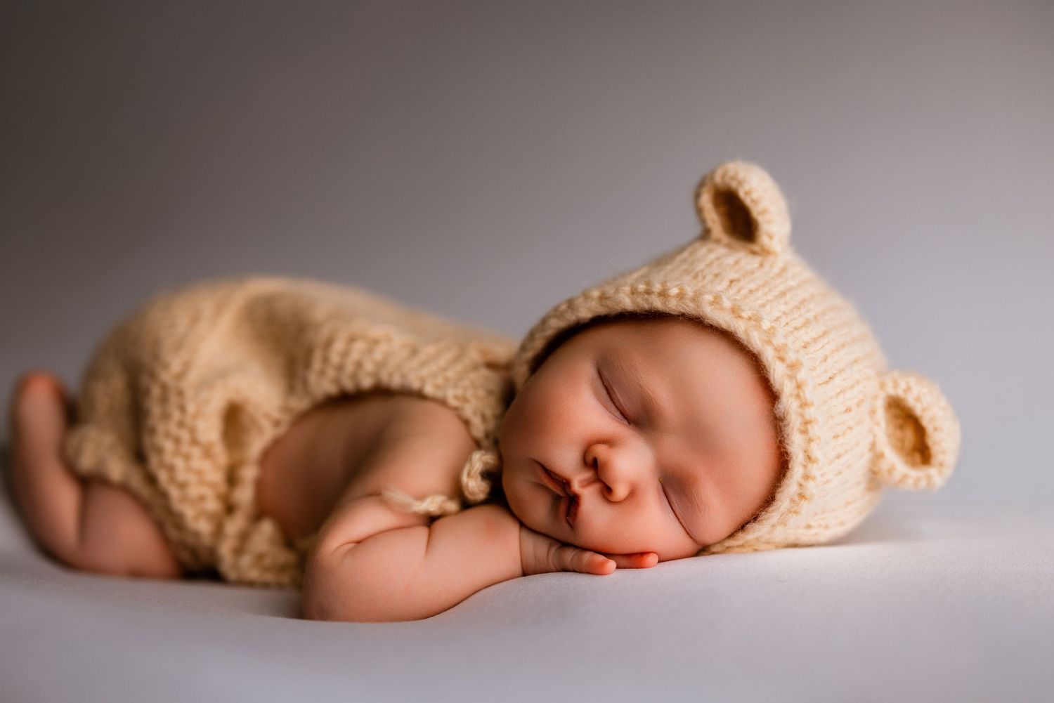 Sleeping newborn baby wearing a cozy cream knitted bear outfit and hat, posed on a white background.
