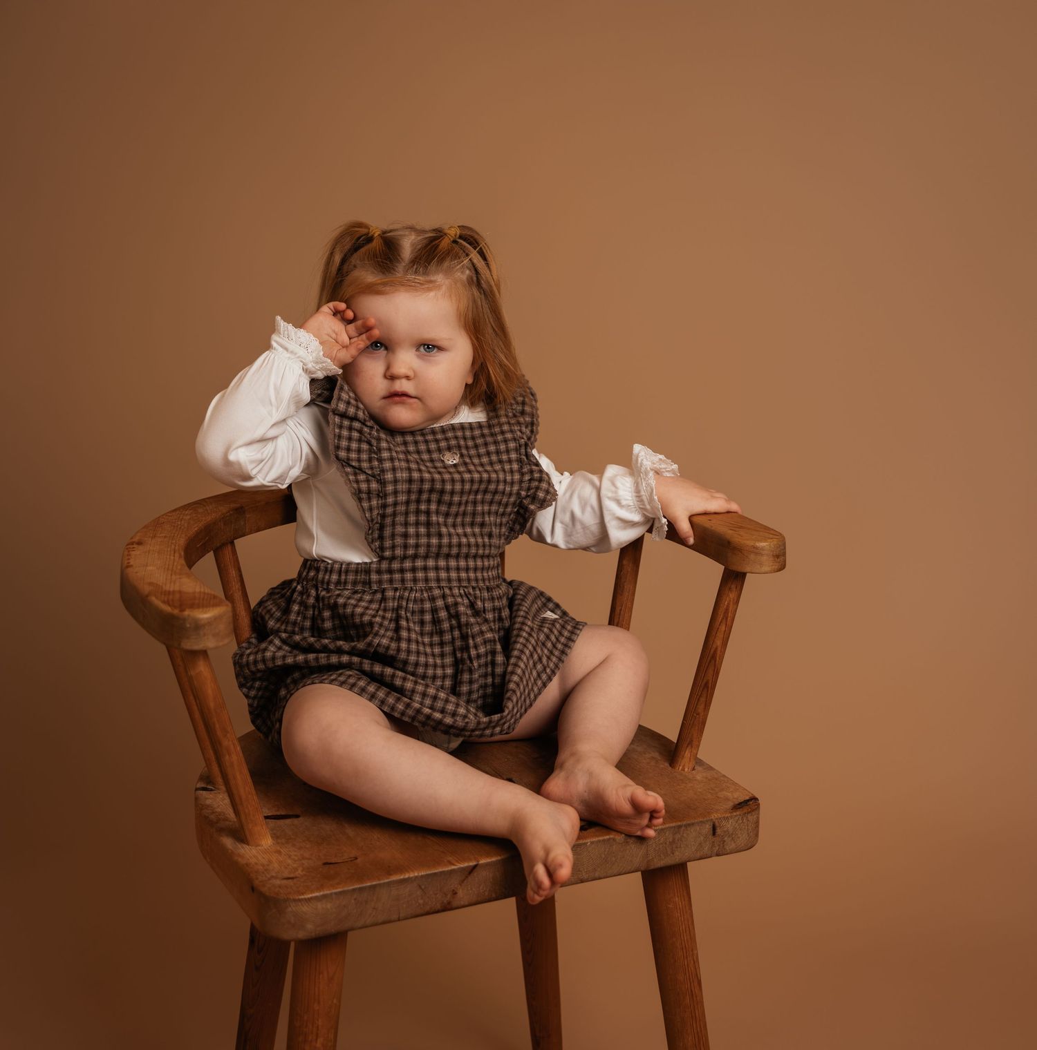 Toddler girl in plaid romper sitting on wooden chair against brown background.