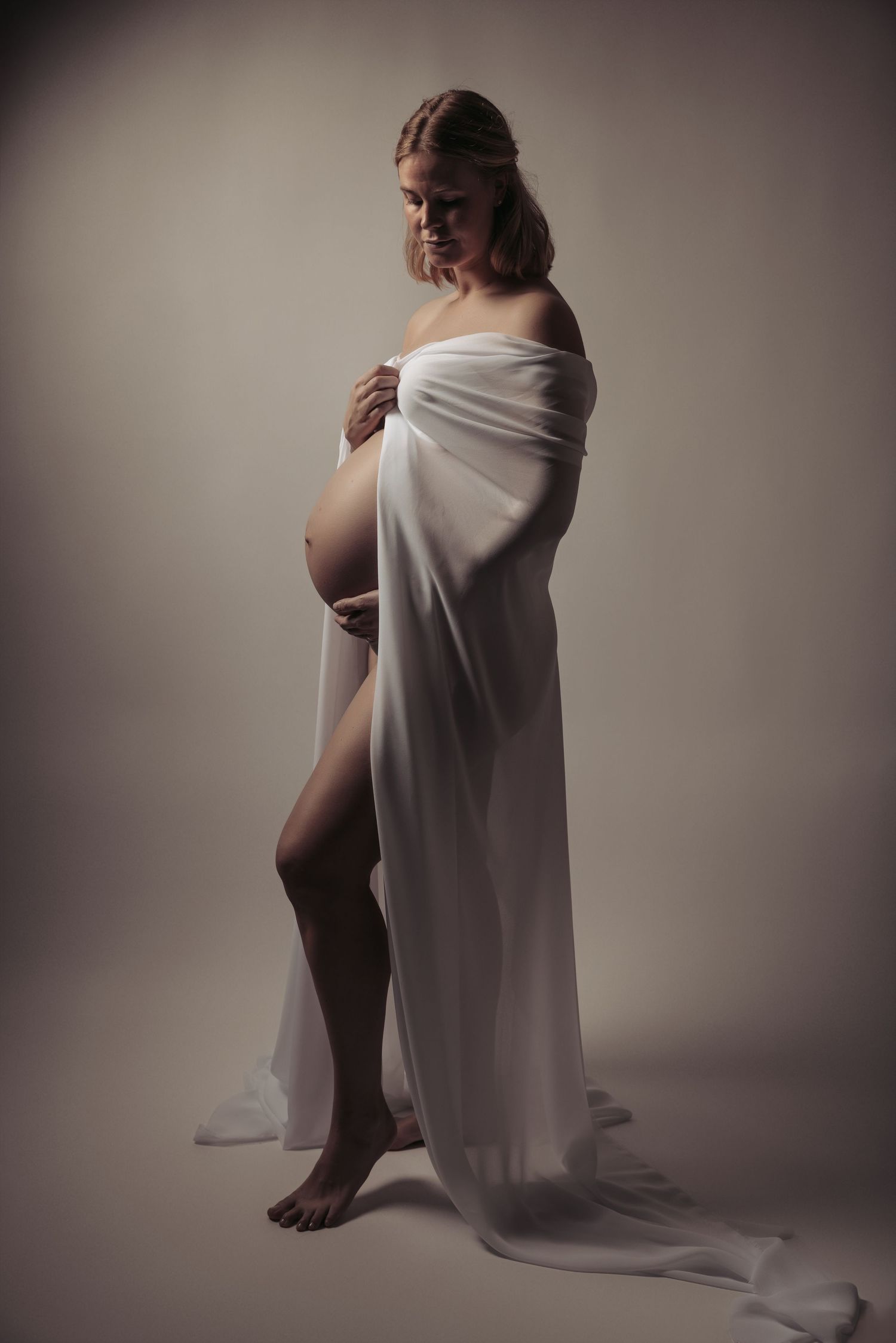 Pregnant woman in sheer white wrap fabric, posing gracefully in soft studio light.