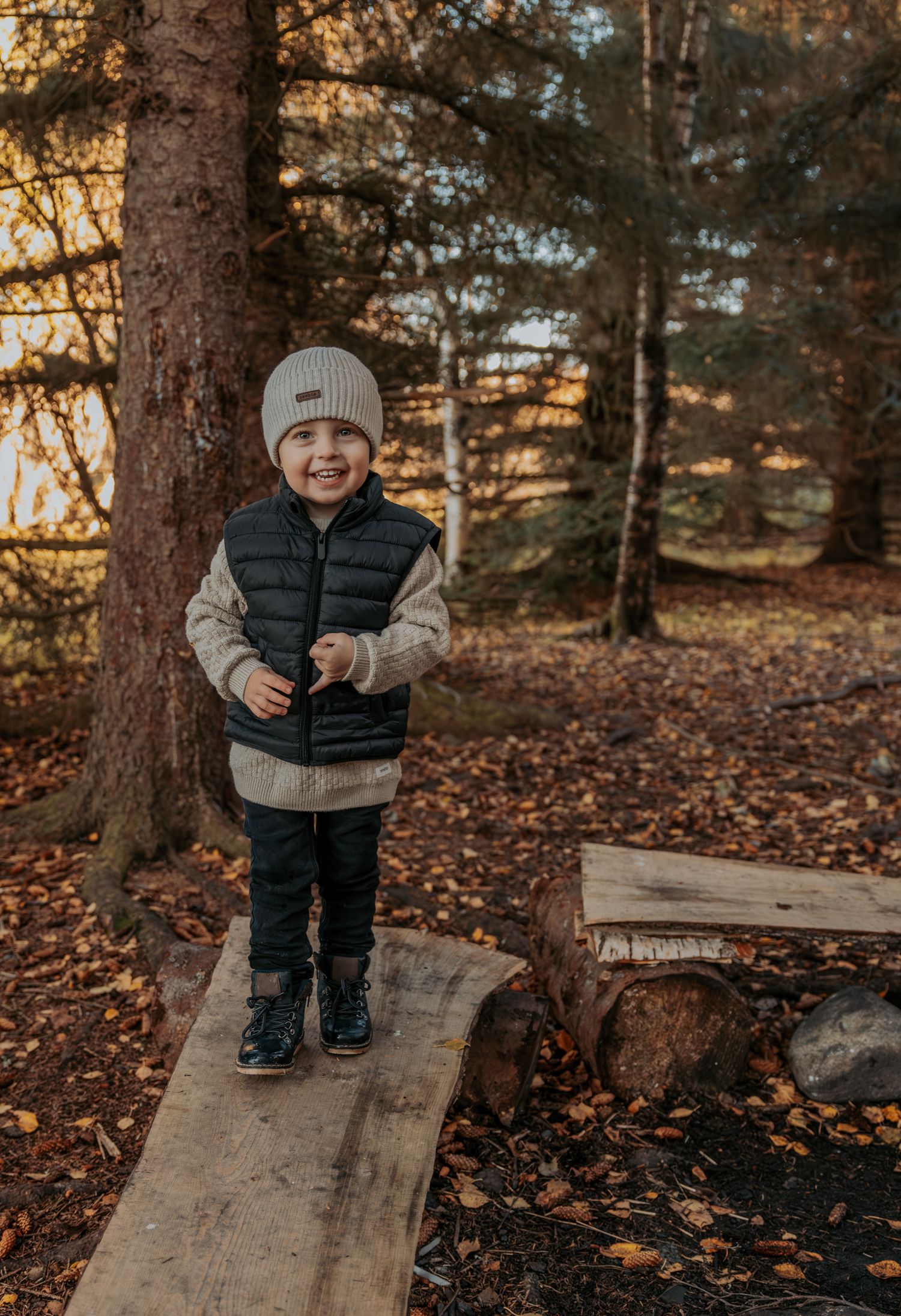 A cheerful toddler in a beanie hat standing on a log in an autumn forest.