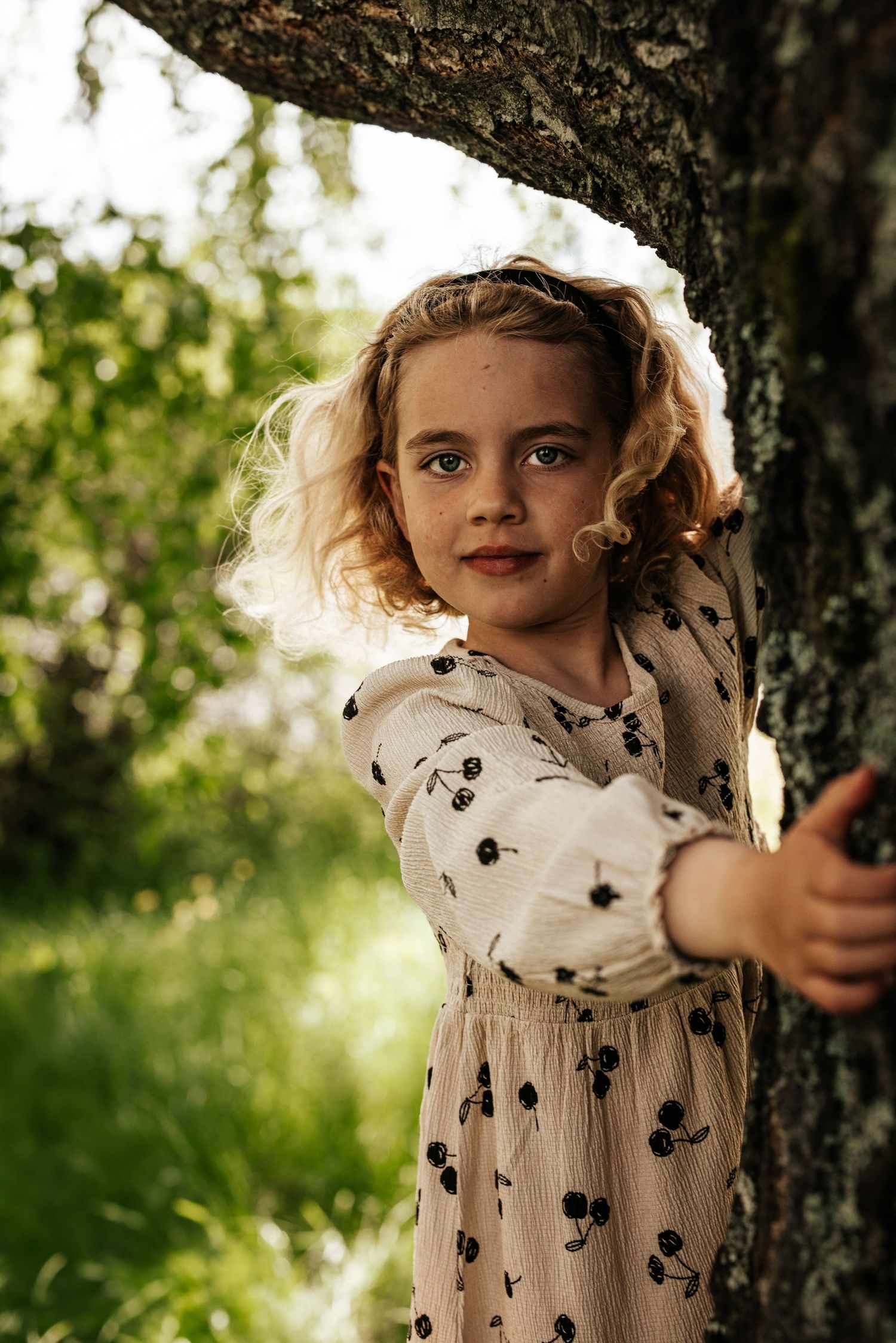 Curly-haired girl in floral dress hugging tree in sunny green garden.