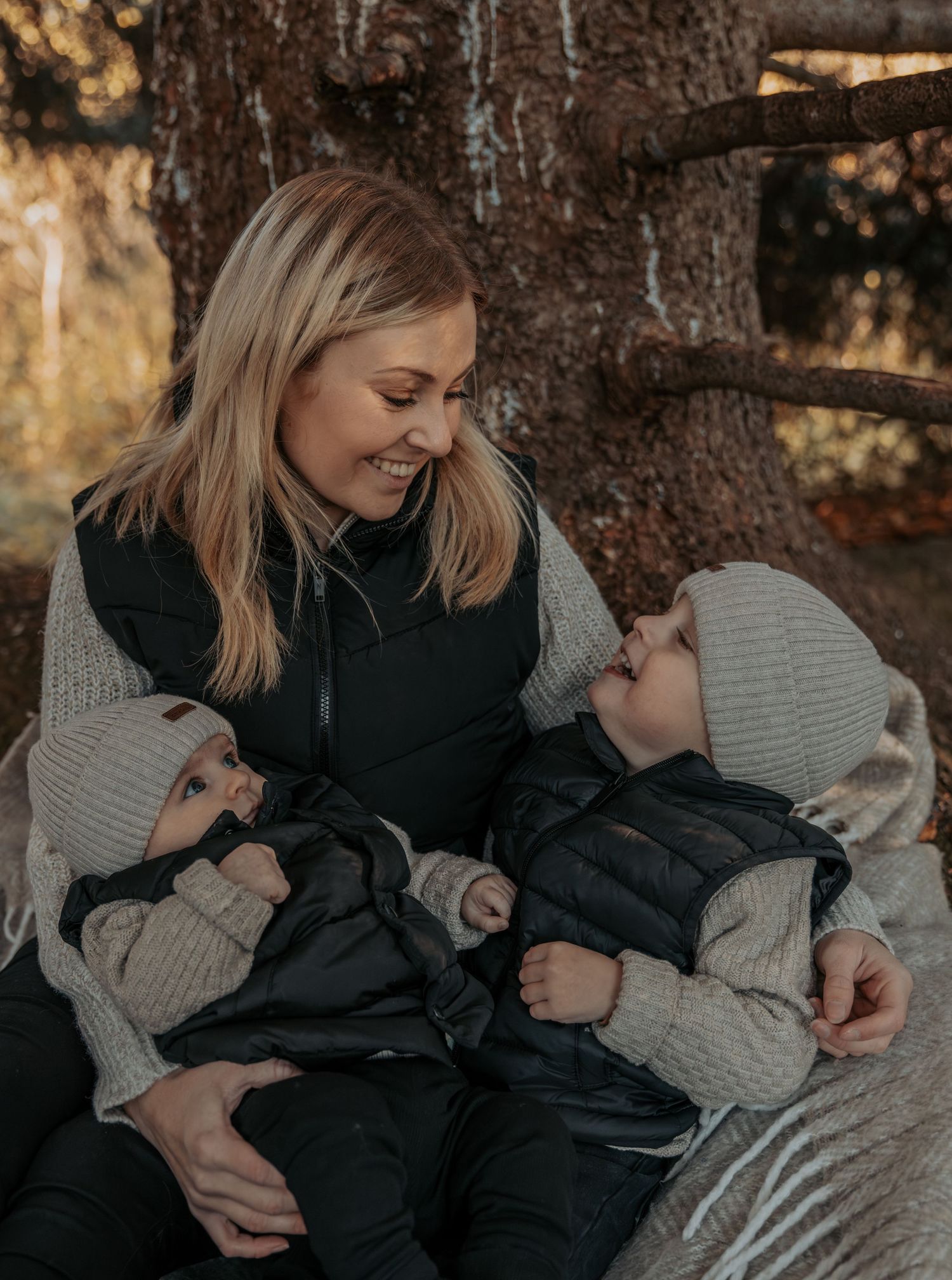 A blonde mother smiling at her two young children in a cozy outdoor autumn setting.