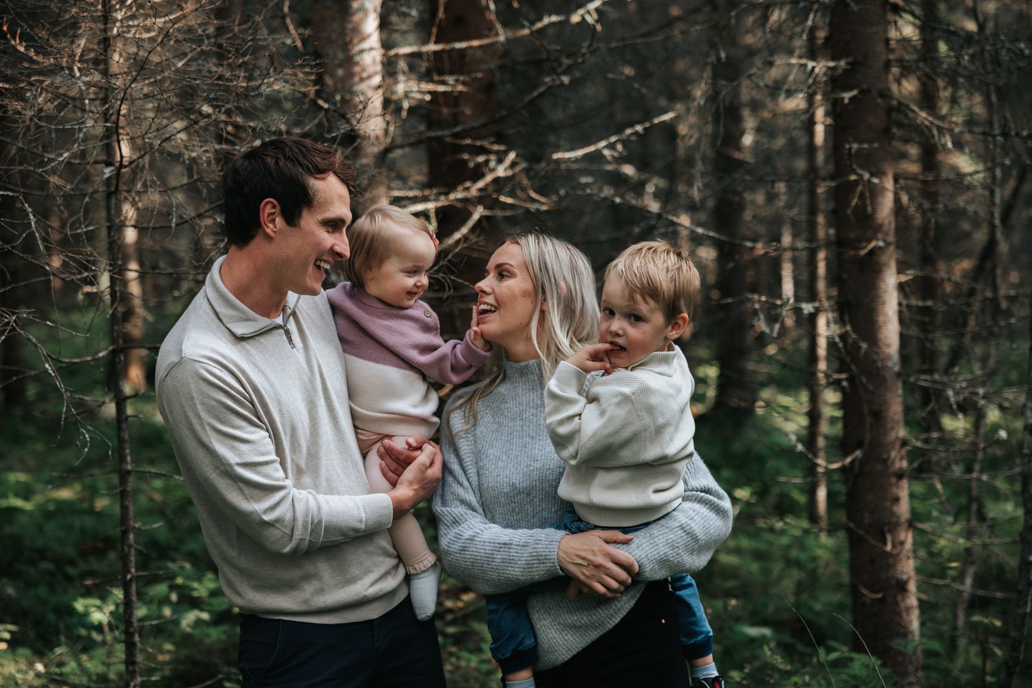 Smiling family of four with two toddlers sharing a joyful moment in a pine forest.