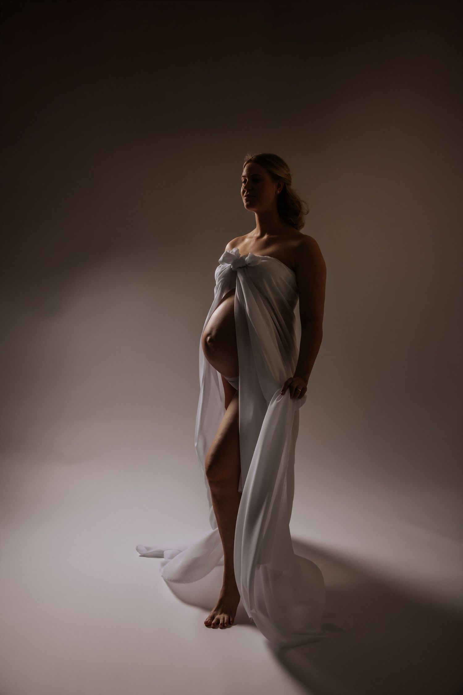 Elegant pregnant woman wrapped in flowing white fabric standing in dramatic studio lighting.