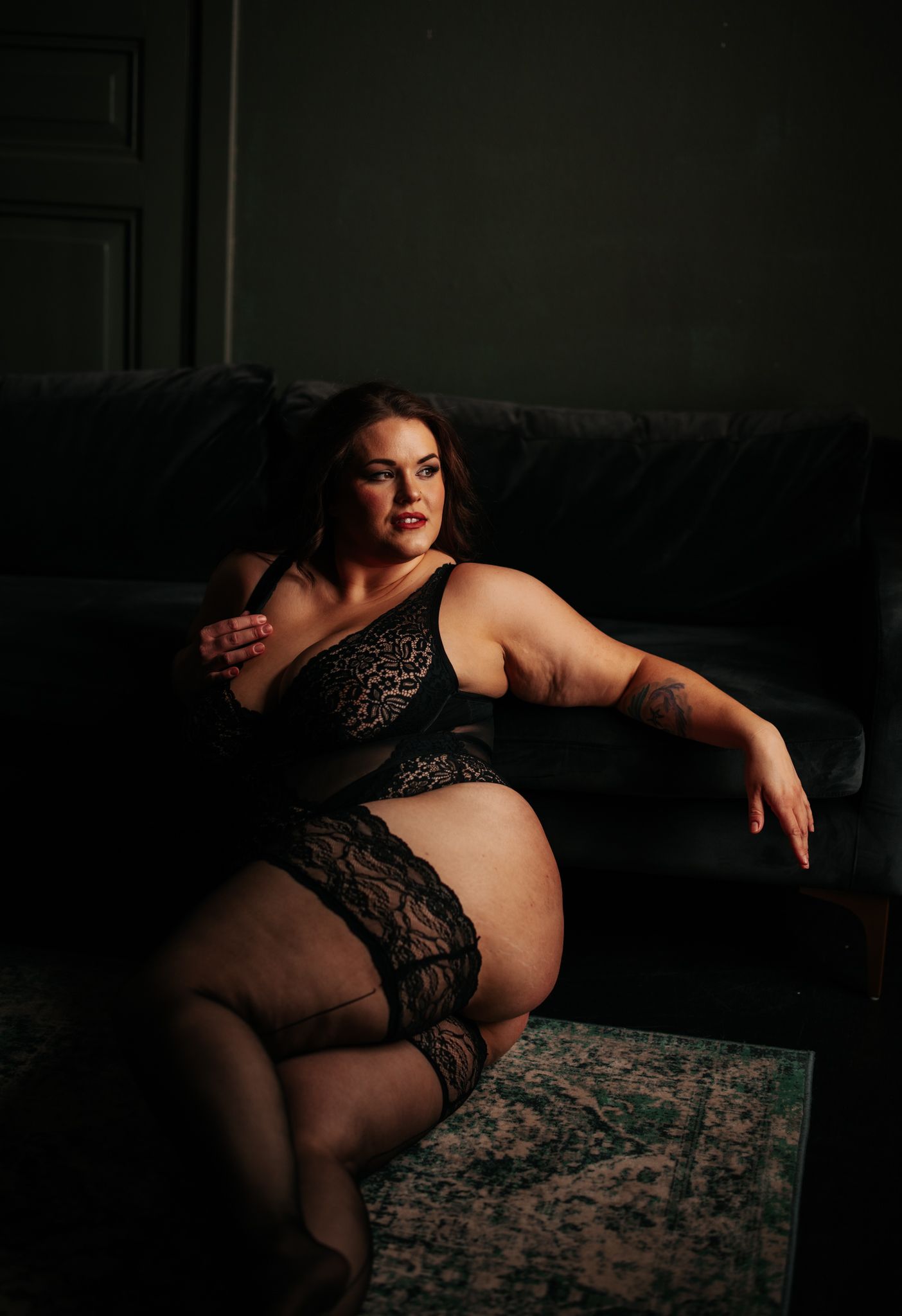 Plus-size woman in black lace lingerie and stockings posing by a dark sofa in boudoir photography.