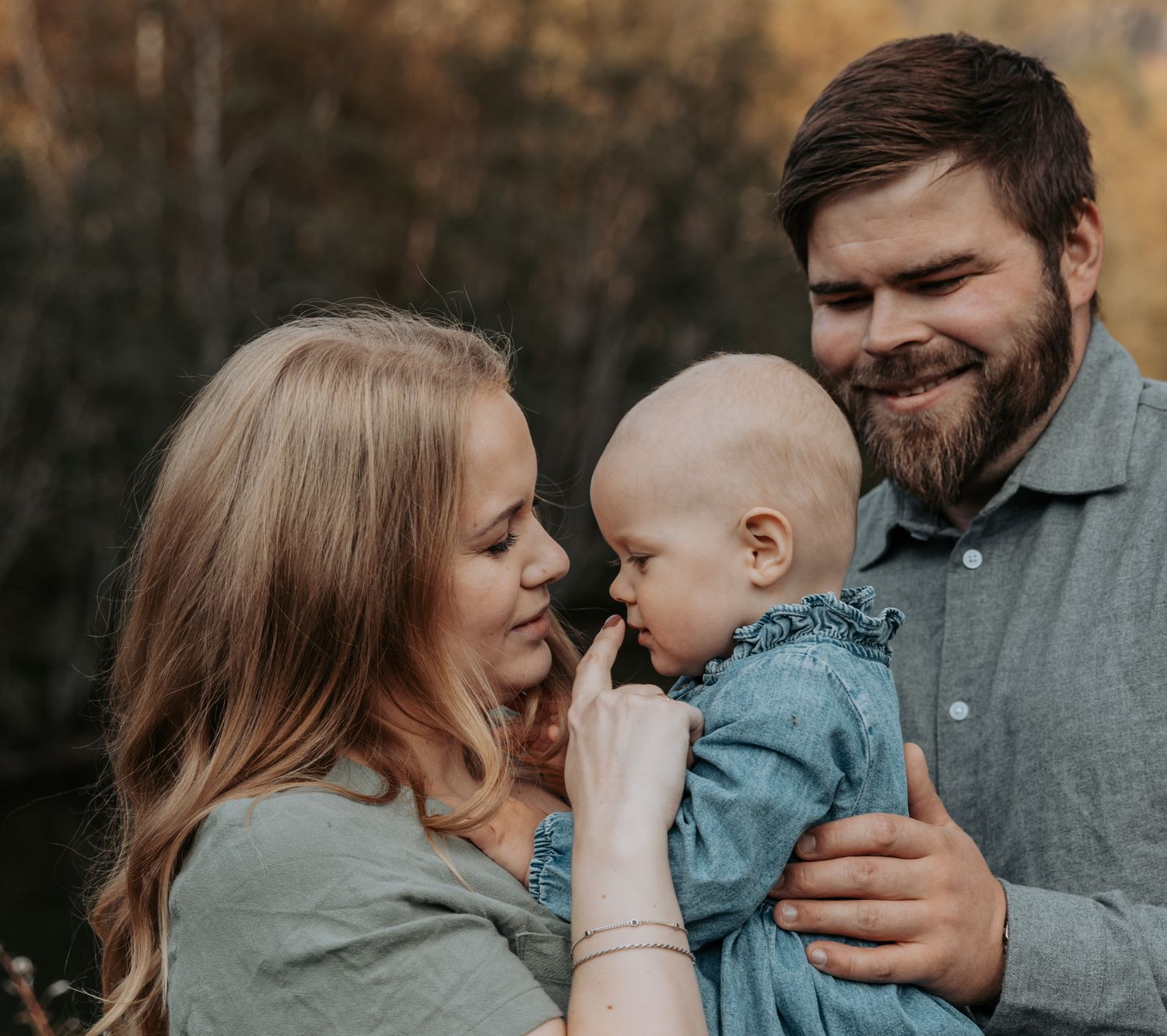 Smiling young family with mother, bearded father, and baby girl posed together in autumn setting.