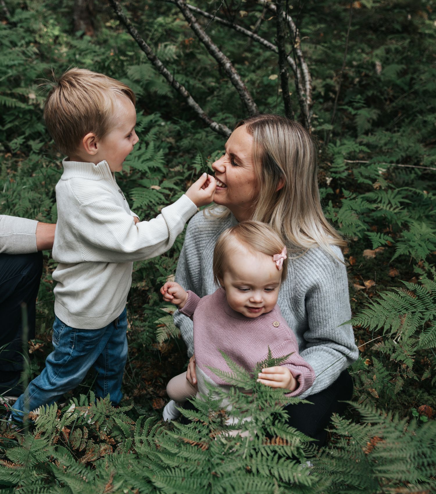 Young boy playfully feeding mother a berry while toddler sister explores forest ferns.