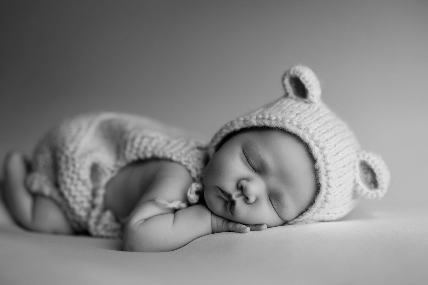 Black and white photo of a sleeping newborn baby wearing a knit bear hat and romper, curled up peacefully.