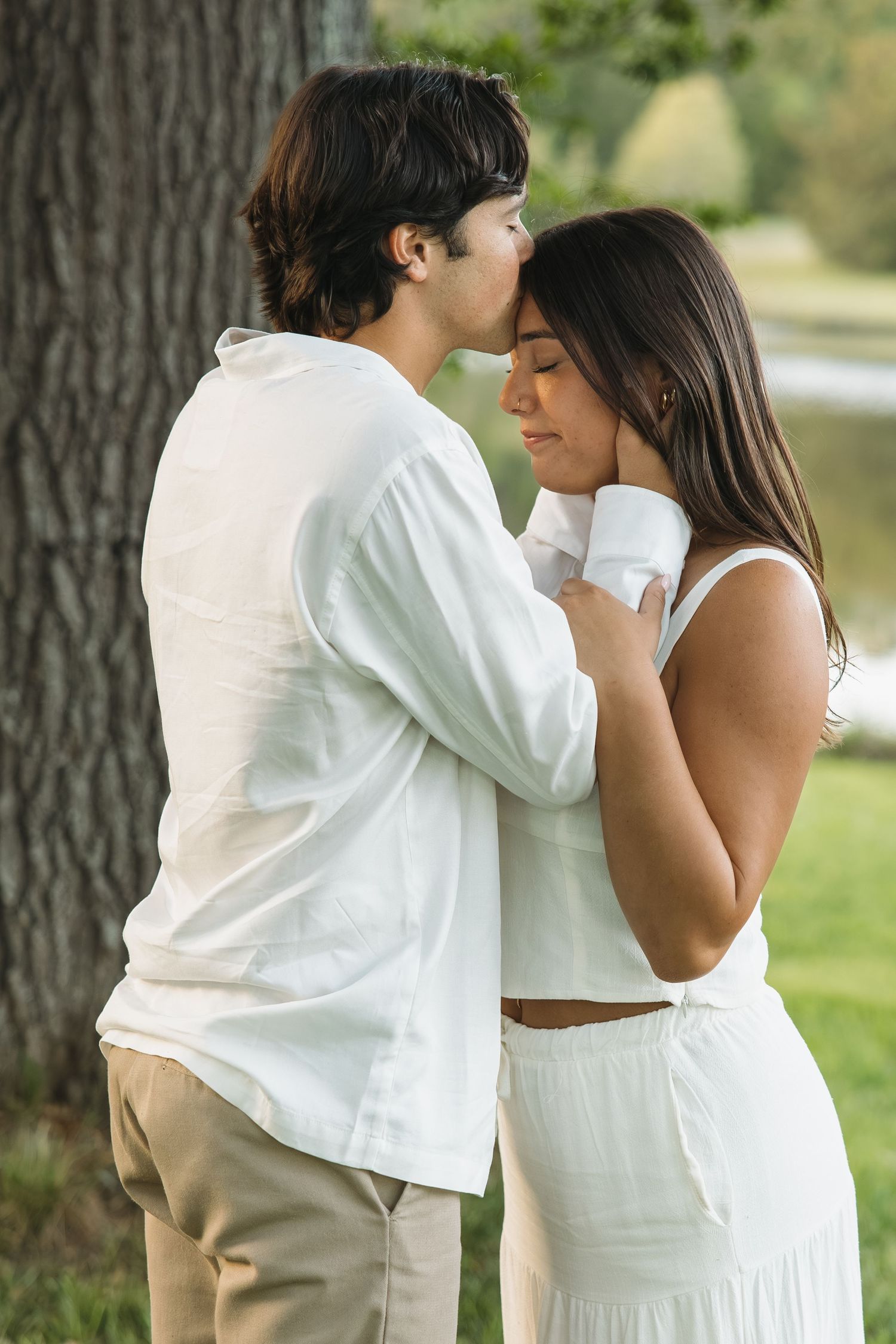 A couple shares an intimate moment, touching foreheads tenderly in a lush green park near a serene lake.