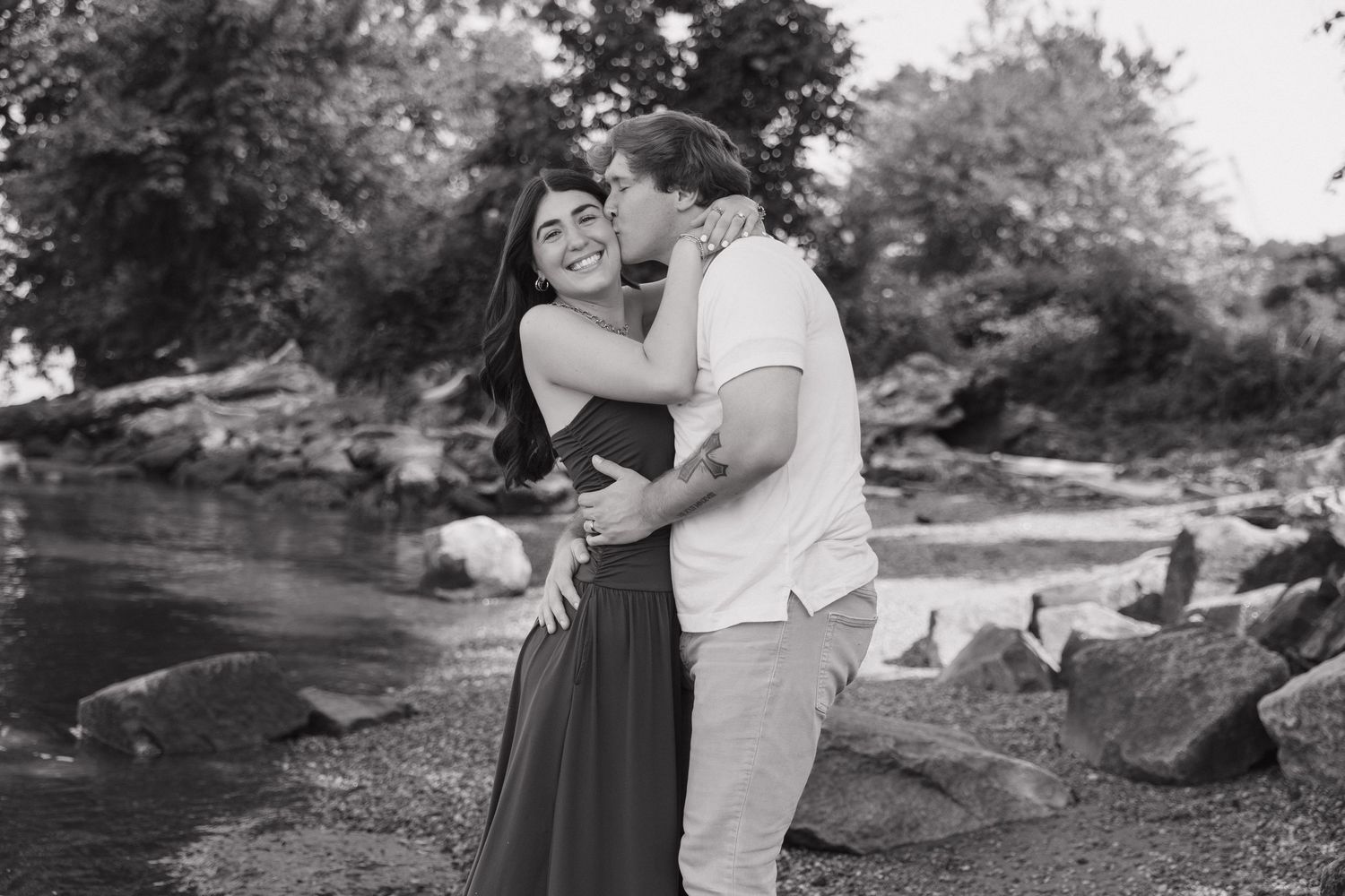 Black and white photo of a couple sharing a tender kiss by a rocky riverbank with trees behind them.