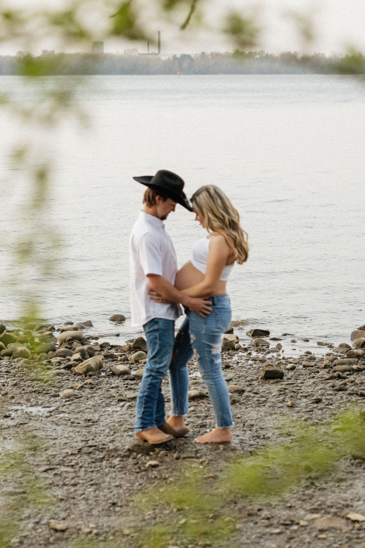 Expectant couple standing forehead to forehead by river, man in black cowboy hat cradling pregnant belly.