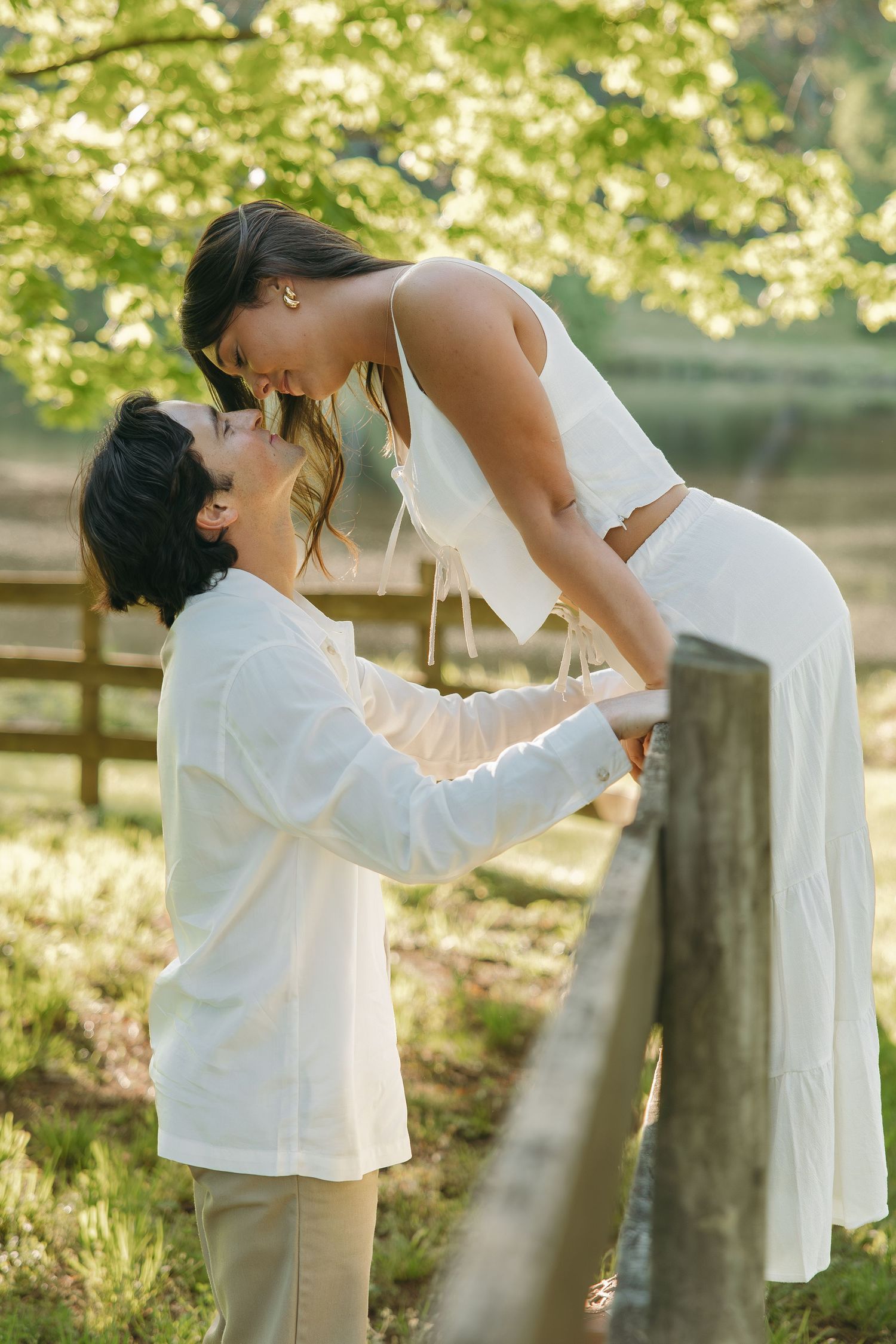 Romantic couple sharing a tender moment on a wooden fence surrounded by lush green trees.