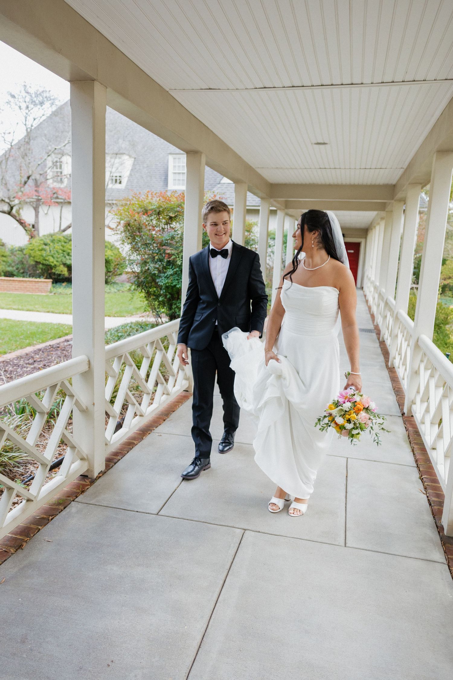 Smiling bride and groom walk together along elegant white columned venue corridor, bride holding colorful bouquet.