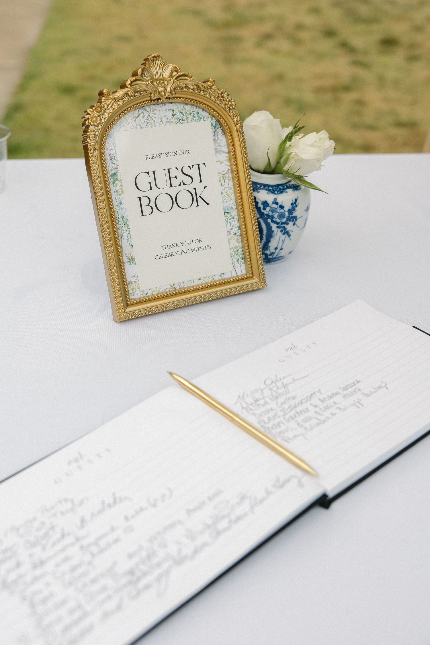 Elegant wedding guest book display with gold frame sign, open journal, gold pen, and white roses.