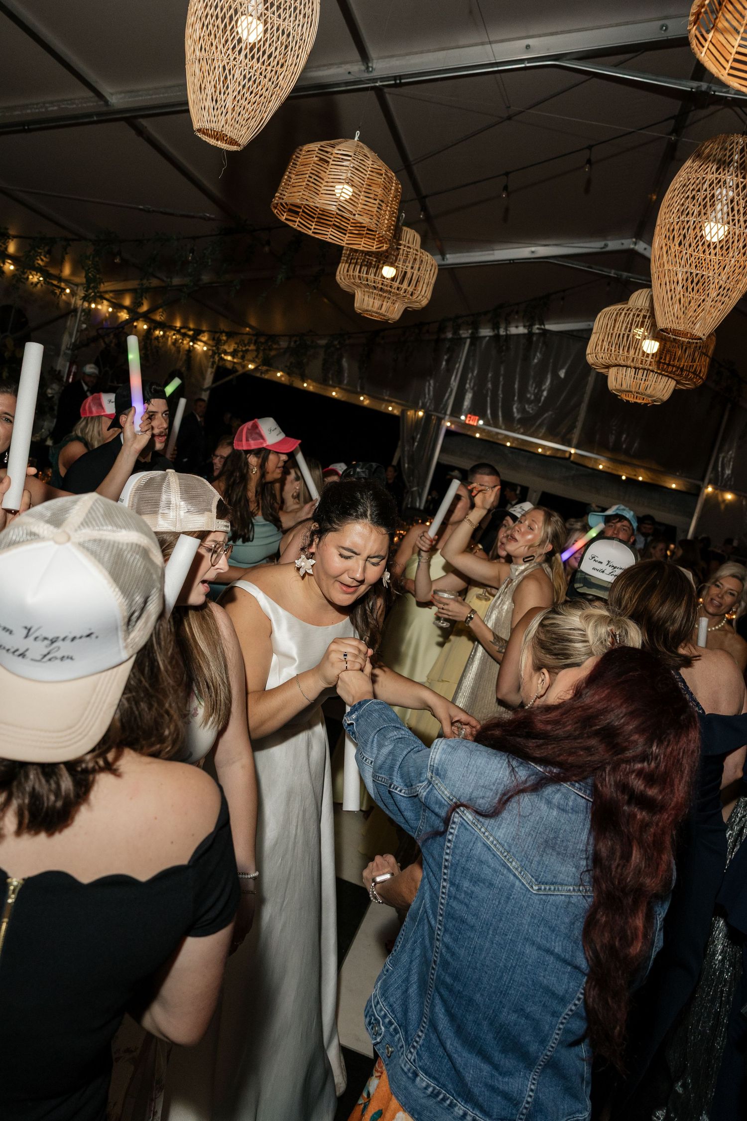 Bride in white gown dancing joyfully with guests at wedding reception under wicker pendant chandeliers.