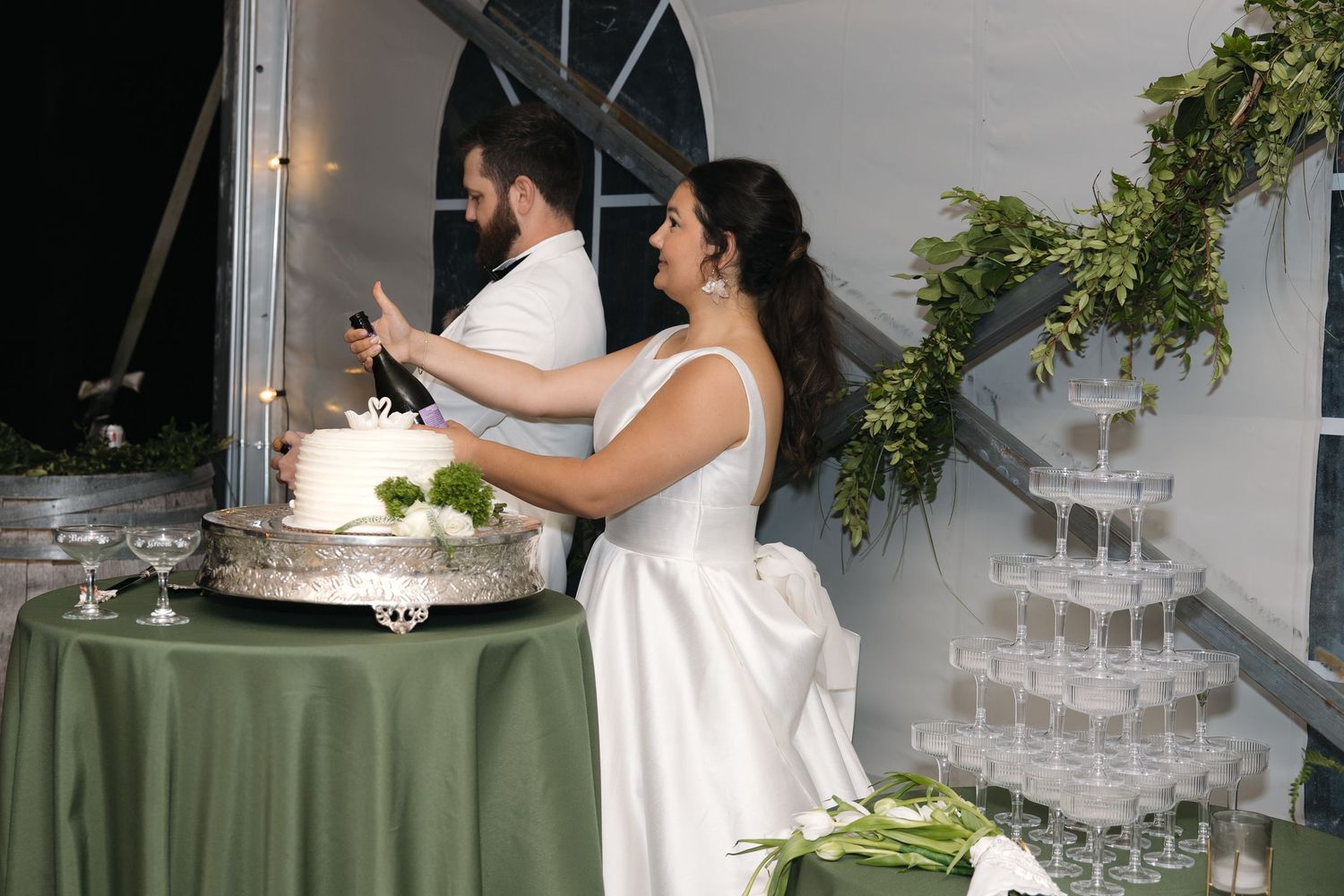 Bride and groom cutting white wedding cake beside champagne tower at tented reception.