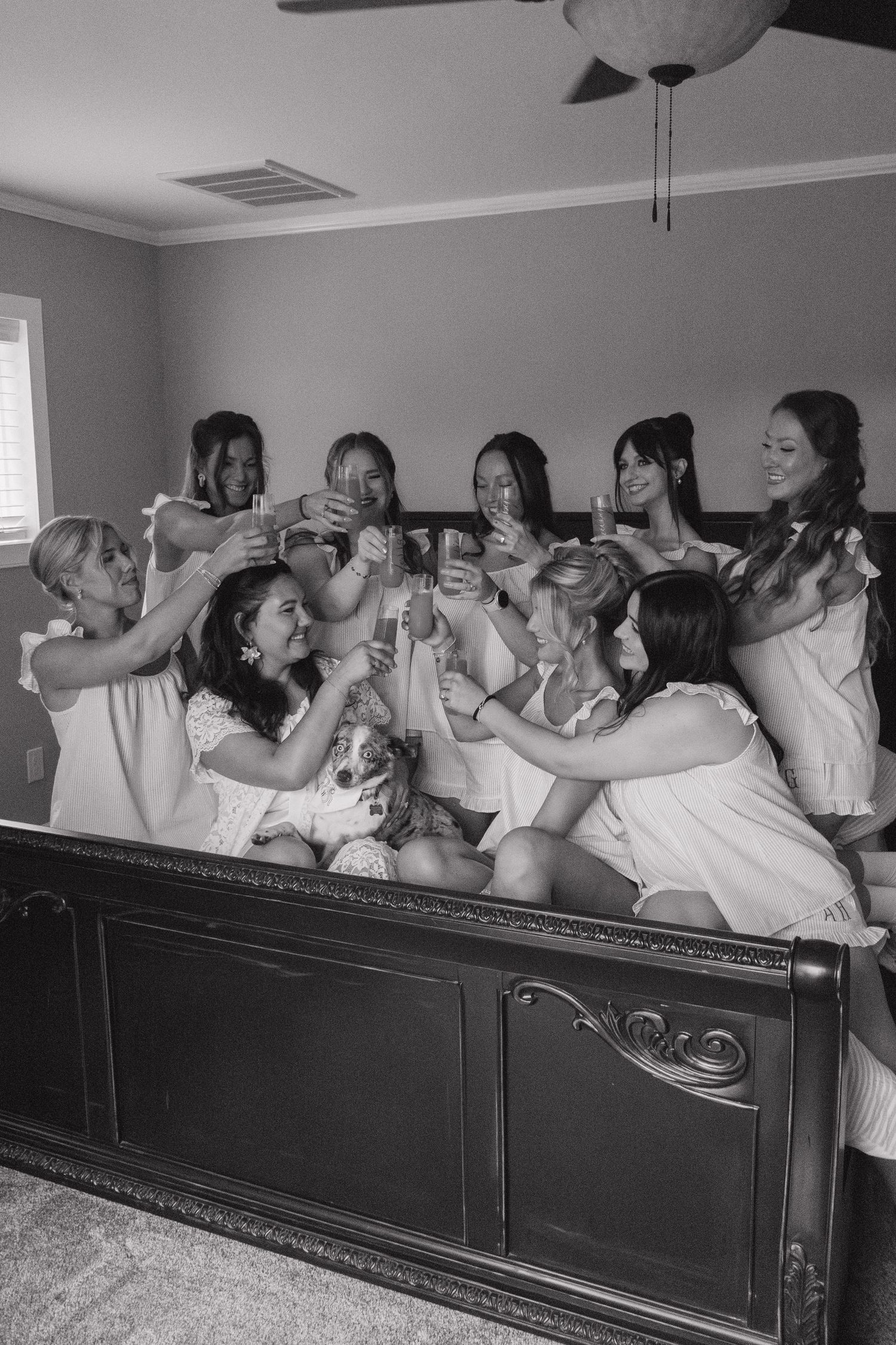 Black and white photo of bridesmaids toasting with champagne glasses on a bed with a dog.